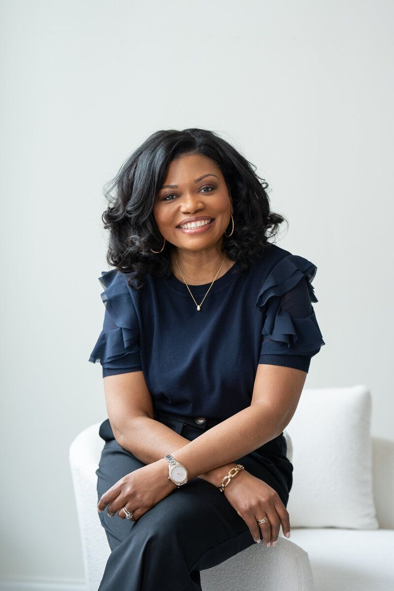 Smiling woman in a navy top seated on a white chair against a light background