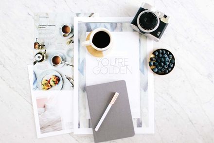Overhead desk flat lay with coffee cups, notebook, pen, magazines, and bowl of blueberries