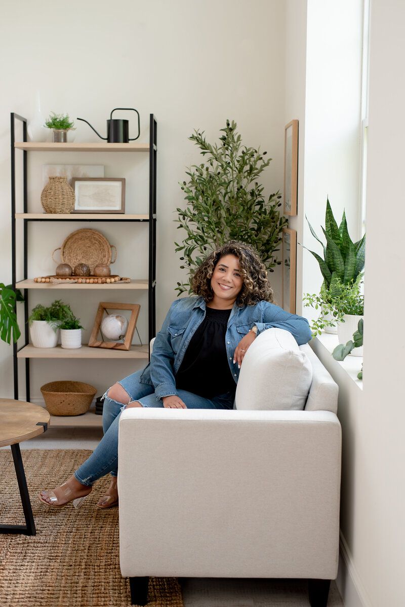 Woman seated on a beige sofa in a bright living room with plants and shelving.