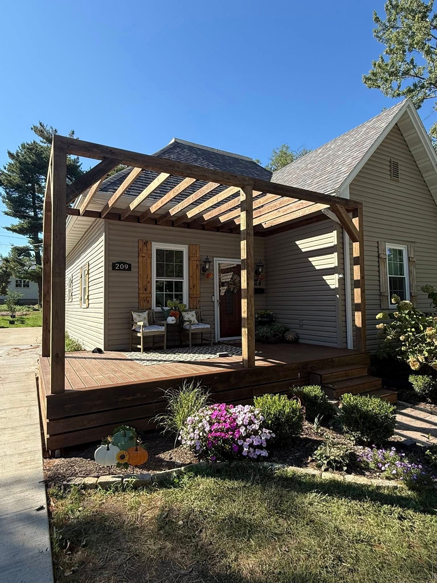 Tan house with wooden pergola over a porch and flower garden in front.