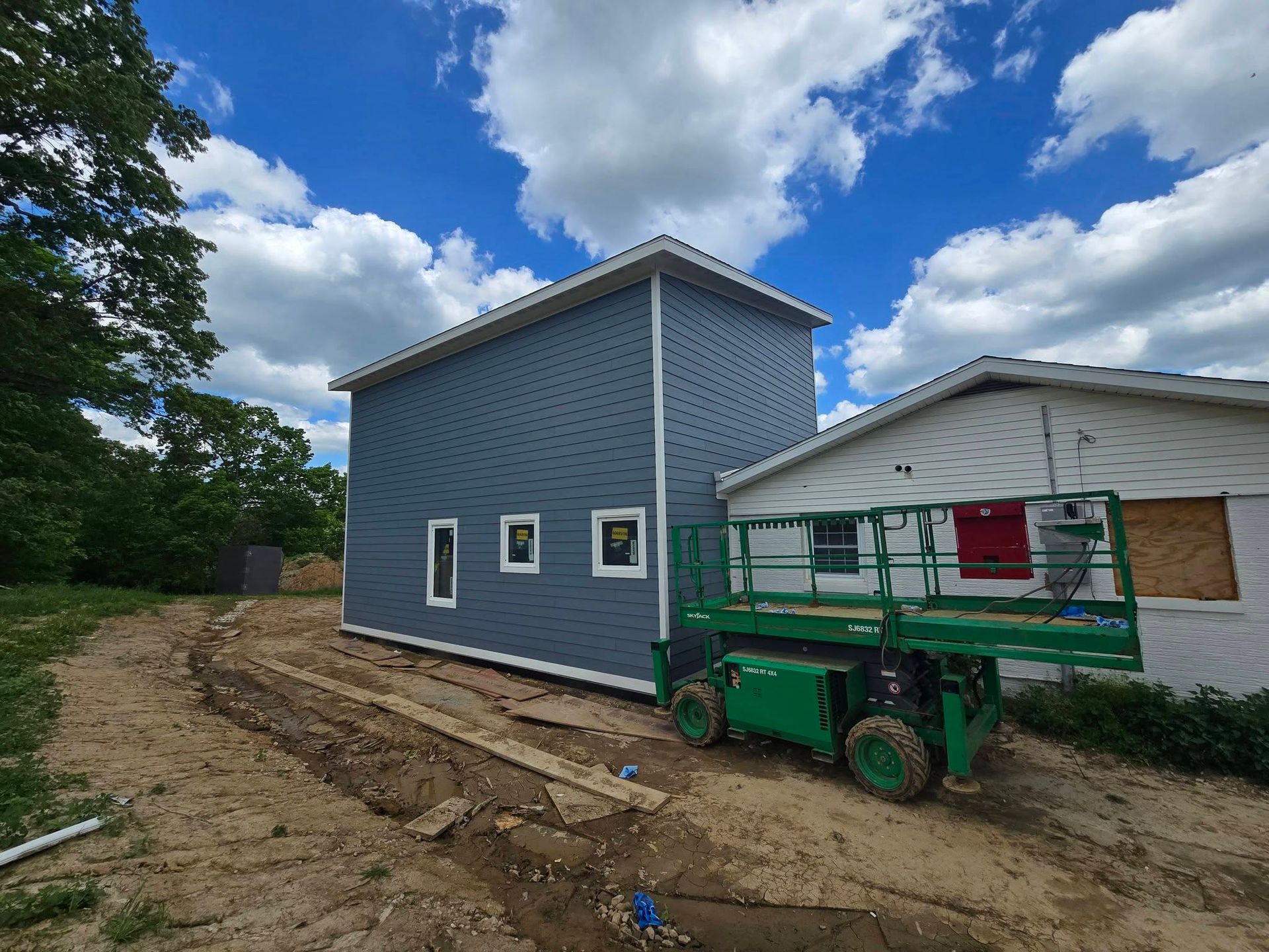 Blue-sided building with small windows being worked on next to a white house, aerial lift present, blue sky.