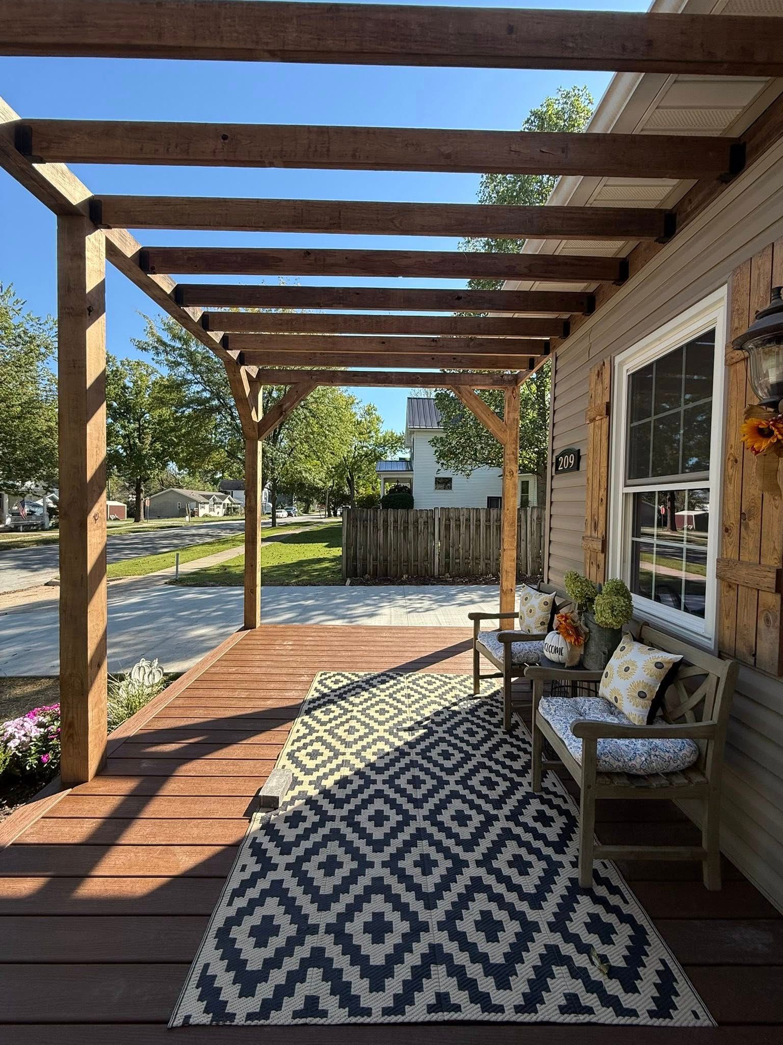 Wooden pergola over a porch with a patterned rug, two chairs, and a window.