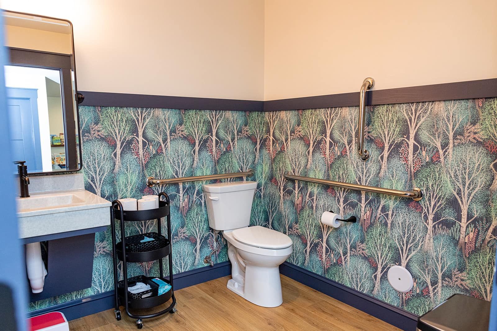 Bathroom with forest wallpaper, accessible toilet with grab bars, small sink, and rolling shelf.
