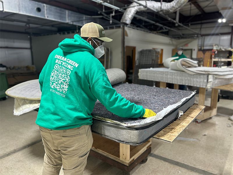 A man in a green hoodie is working on a mattress in a factory.