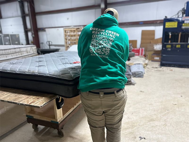 A man in a green hoodie is standing next to a mattress in a warehouse.