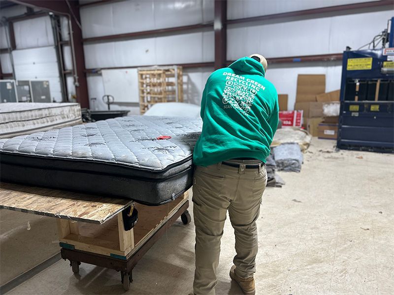 A man in a green hoodie is standing next to a mattress in a warehouse.