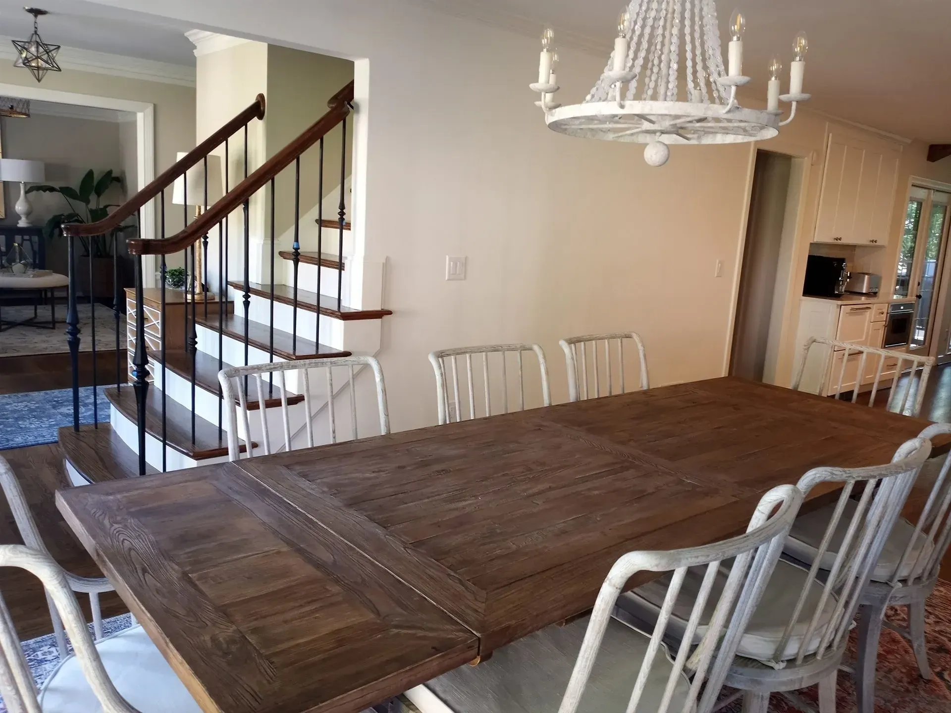 Dining room with large wooden table and white chairs, stairs visible in the background.