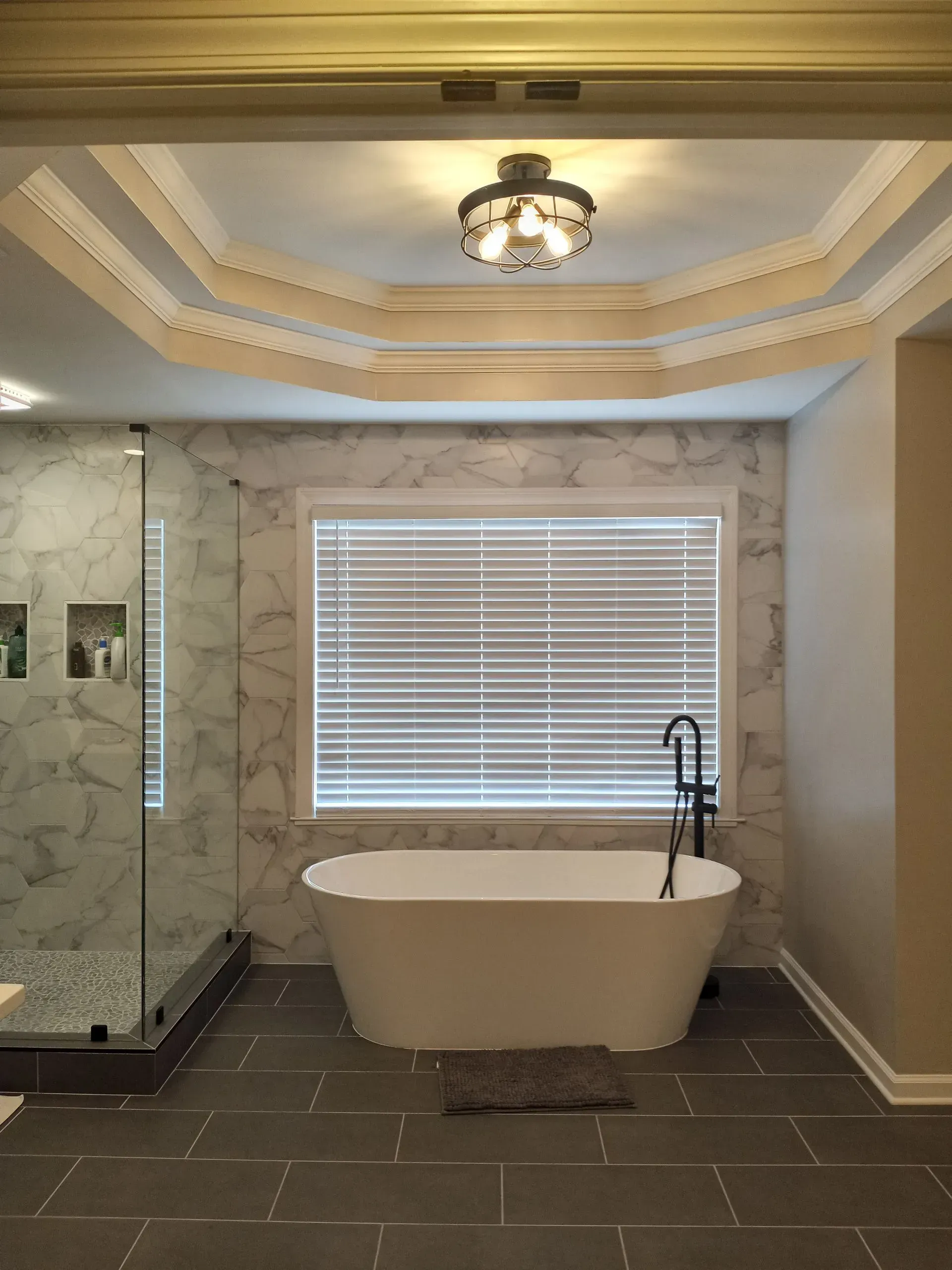 Bathroom with a white freestanding tub, large window with blinds, marble tile walls, and decorative ceiling trim.