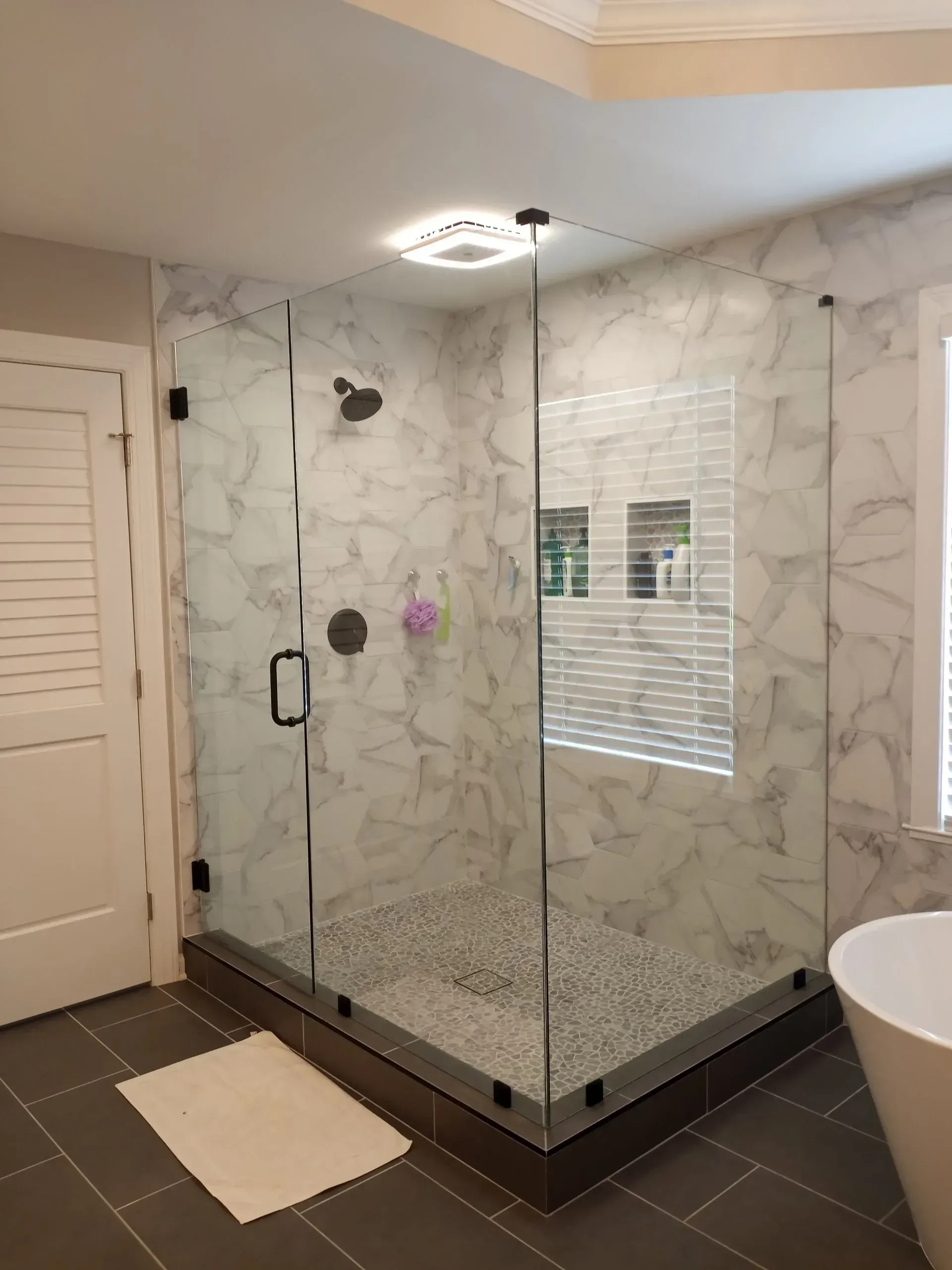 Modern glass shower with white marble-look tile, pebble floor, and dark hardware. Adjacent to a door and a window.