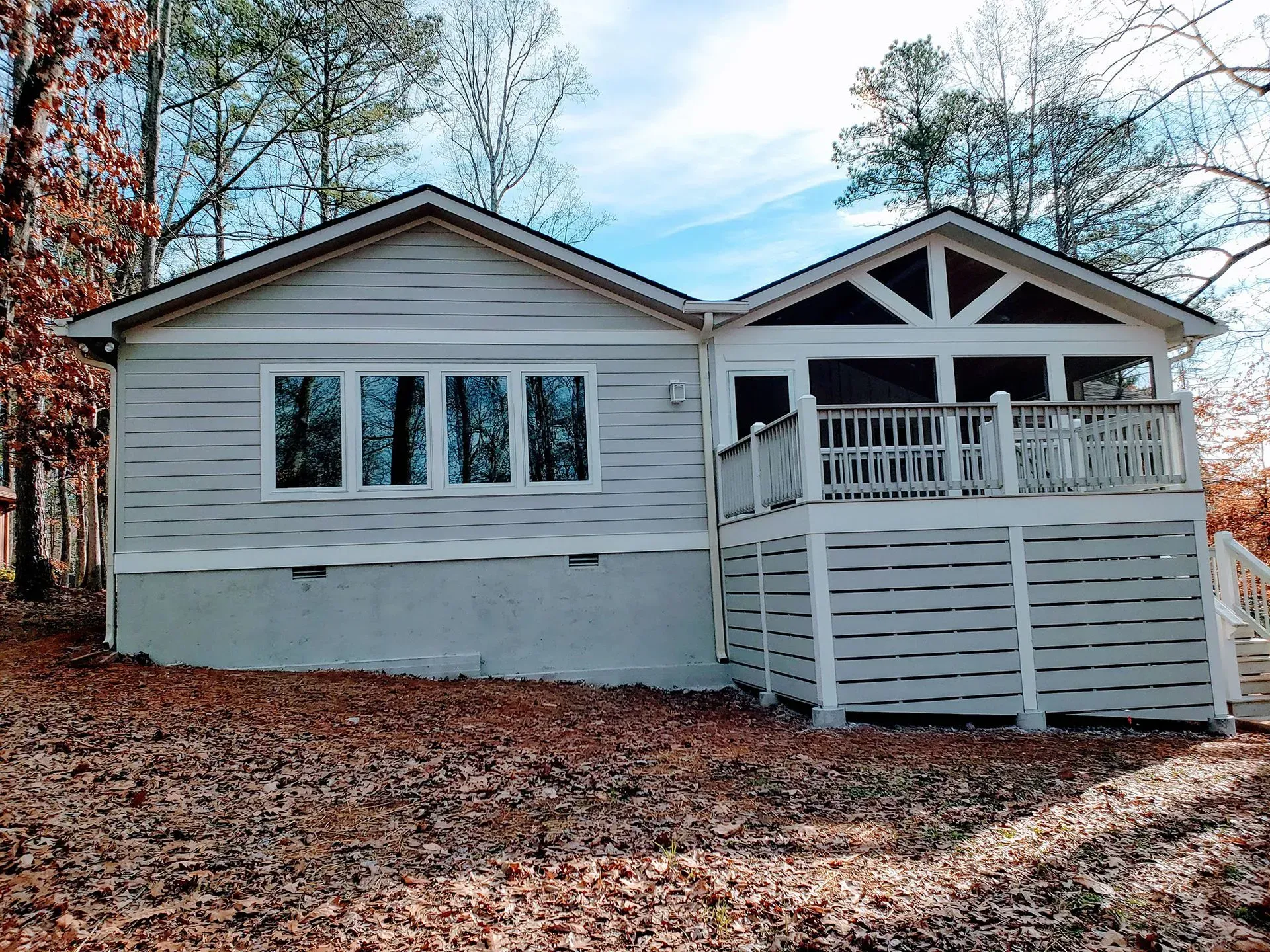Cottage with light siding and a deck with horizontal railings surrounded by trees.