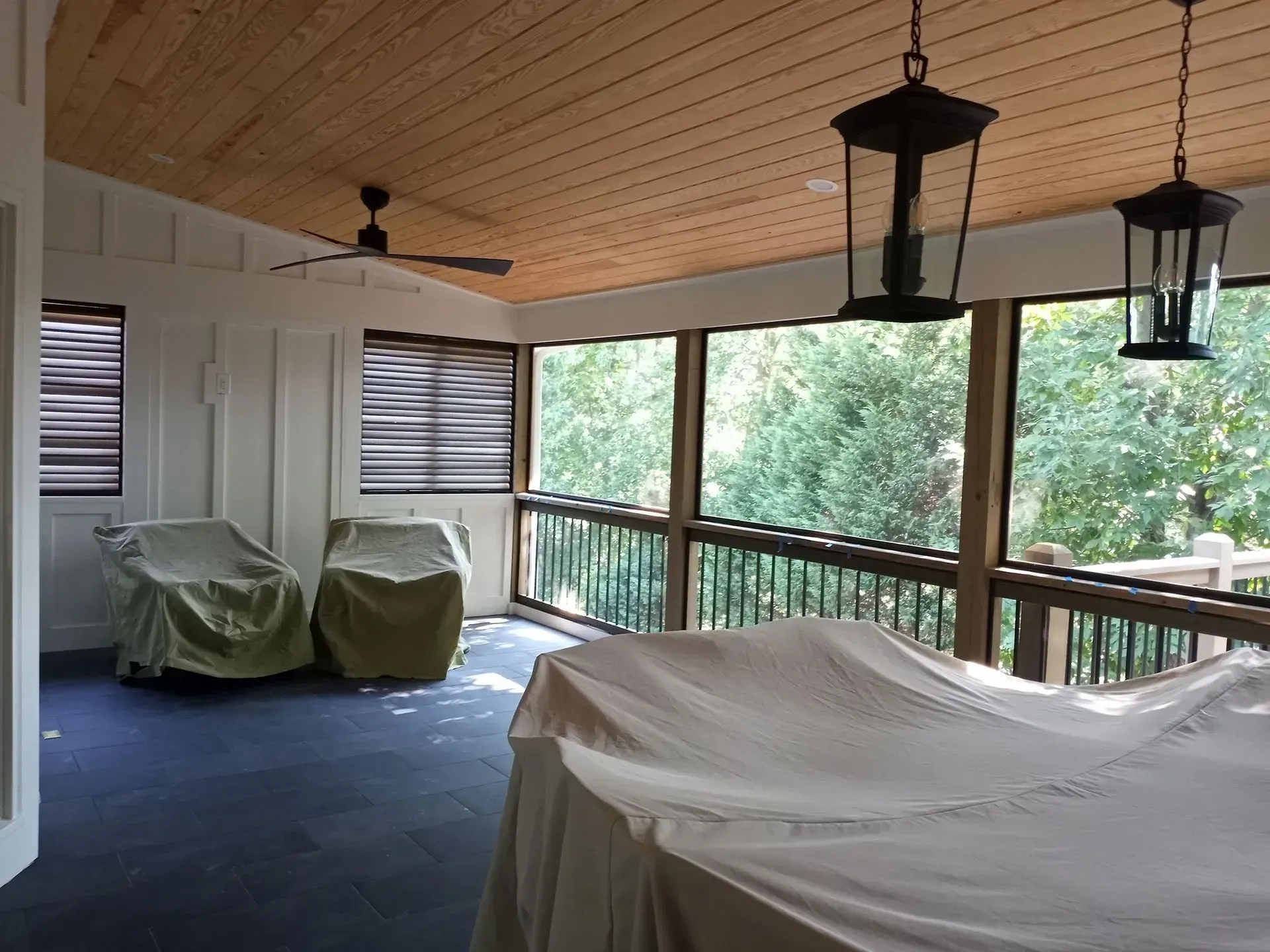 Screened porch with covered furniture, wood ceiling, dark trim, and a view of trees.
