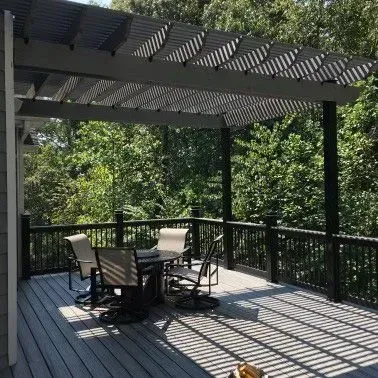 Deck with pergola: gray deck, black railing, outdoor table and chairs, wooded background.