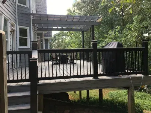 Deck with black railing, pergola, and grill, set against a backdrop of trees and a house with grey siding.