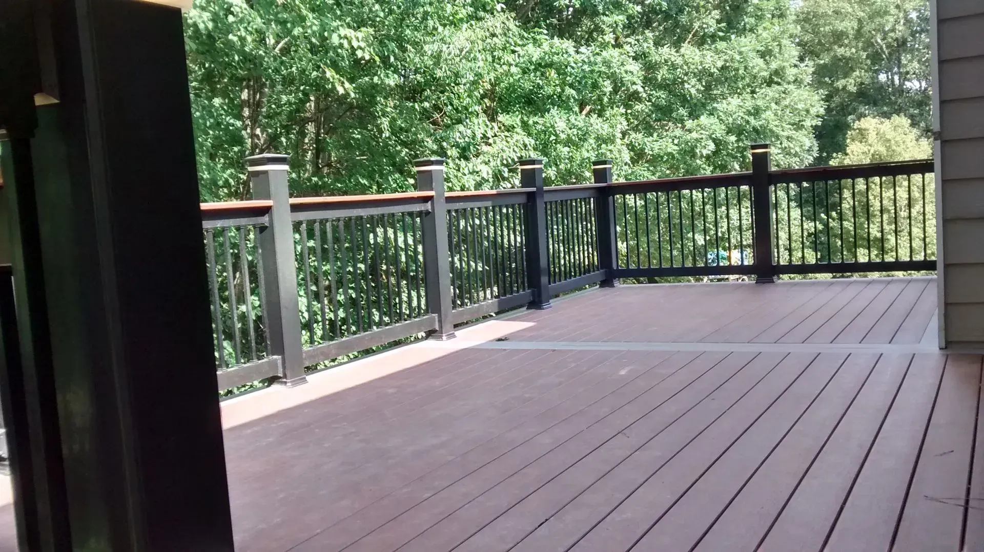 Deck with brown composite decking, black railing, and lush green trees in the background.