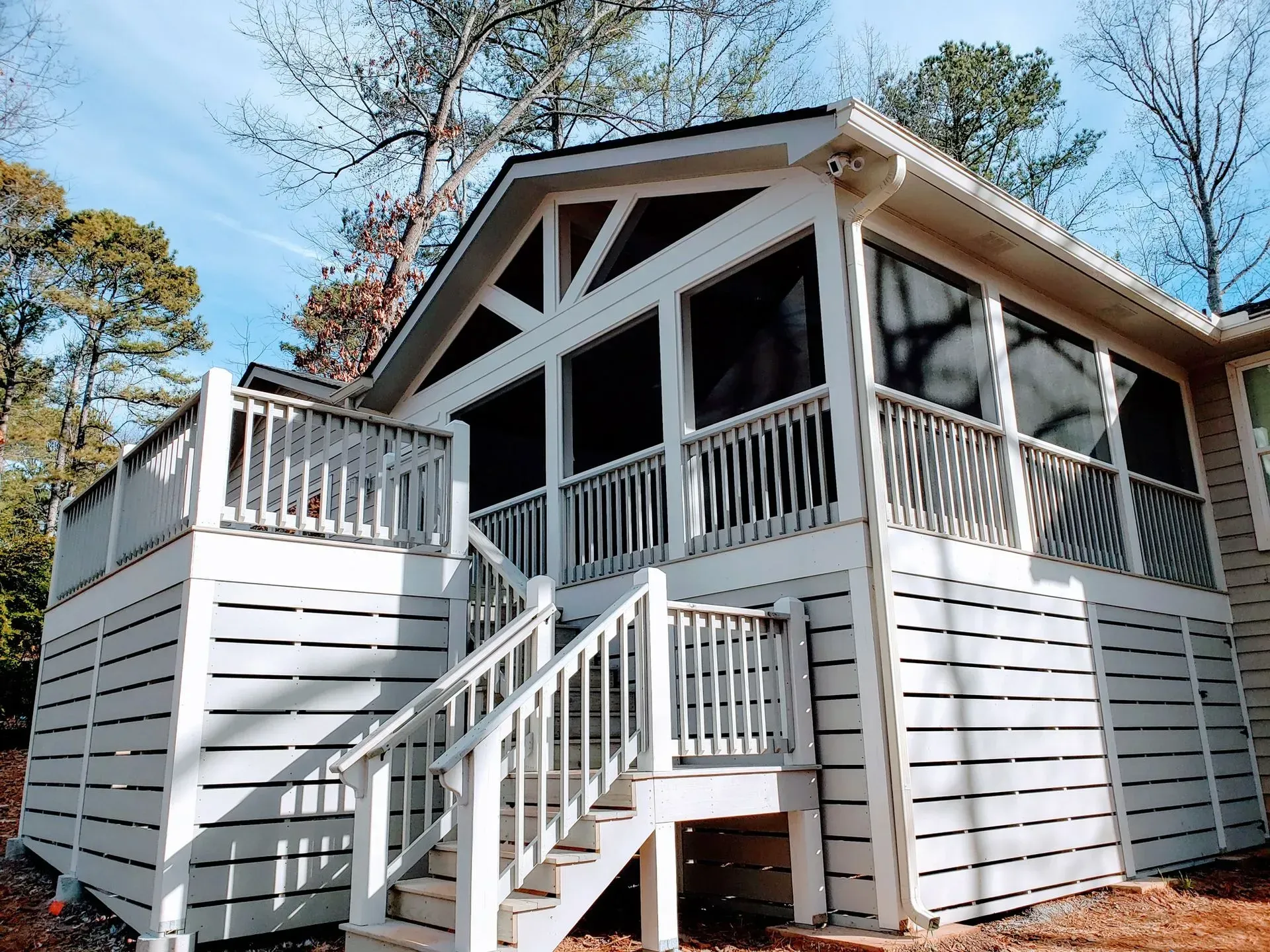 White screened-in porch with deck and staircase, attached to a house; surrounded by trees.