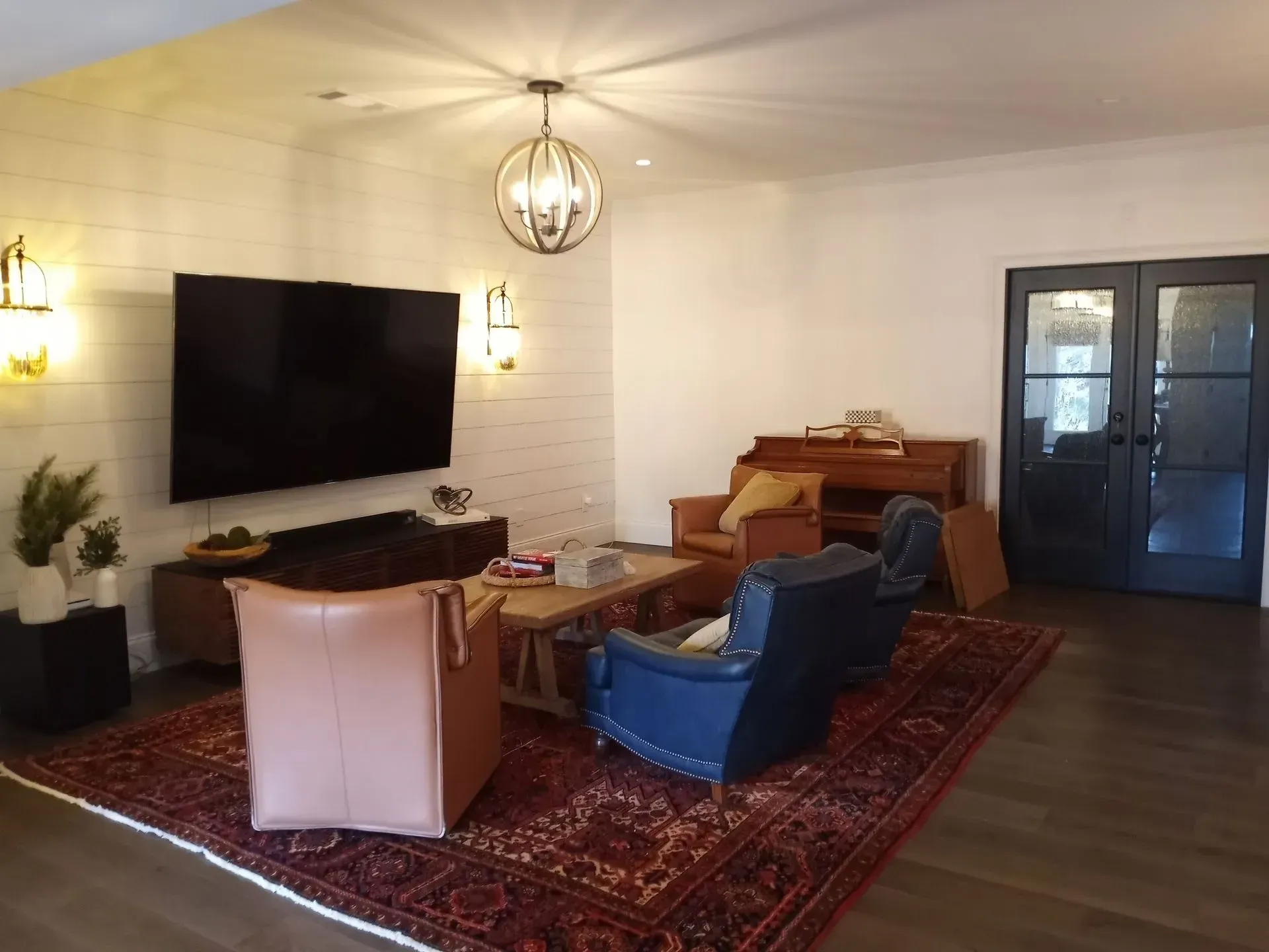Living room: TV on brick wall, leather chairs on patterned rug, piano, glass door.