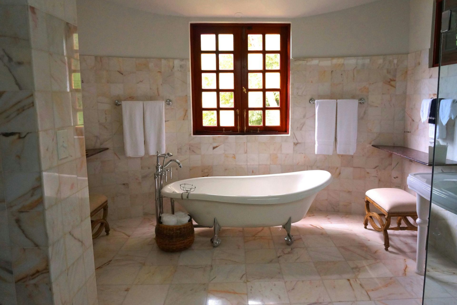 Bathroom with claw-foot tub, wood-framed window, and marble tile walls and floor. Towels hang.