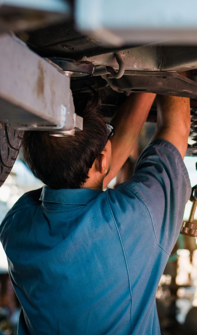 Mechanic working on a car, wearing blue uniform, seen from behind, underneath the vehicle.
