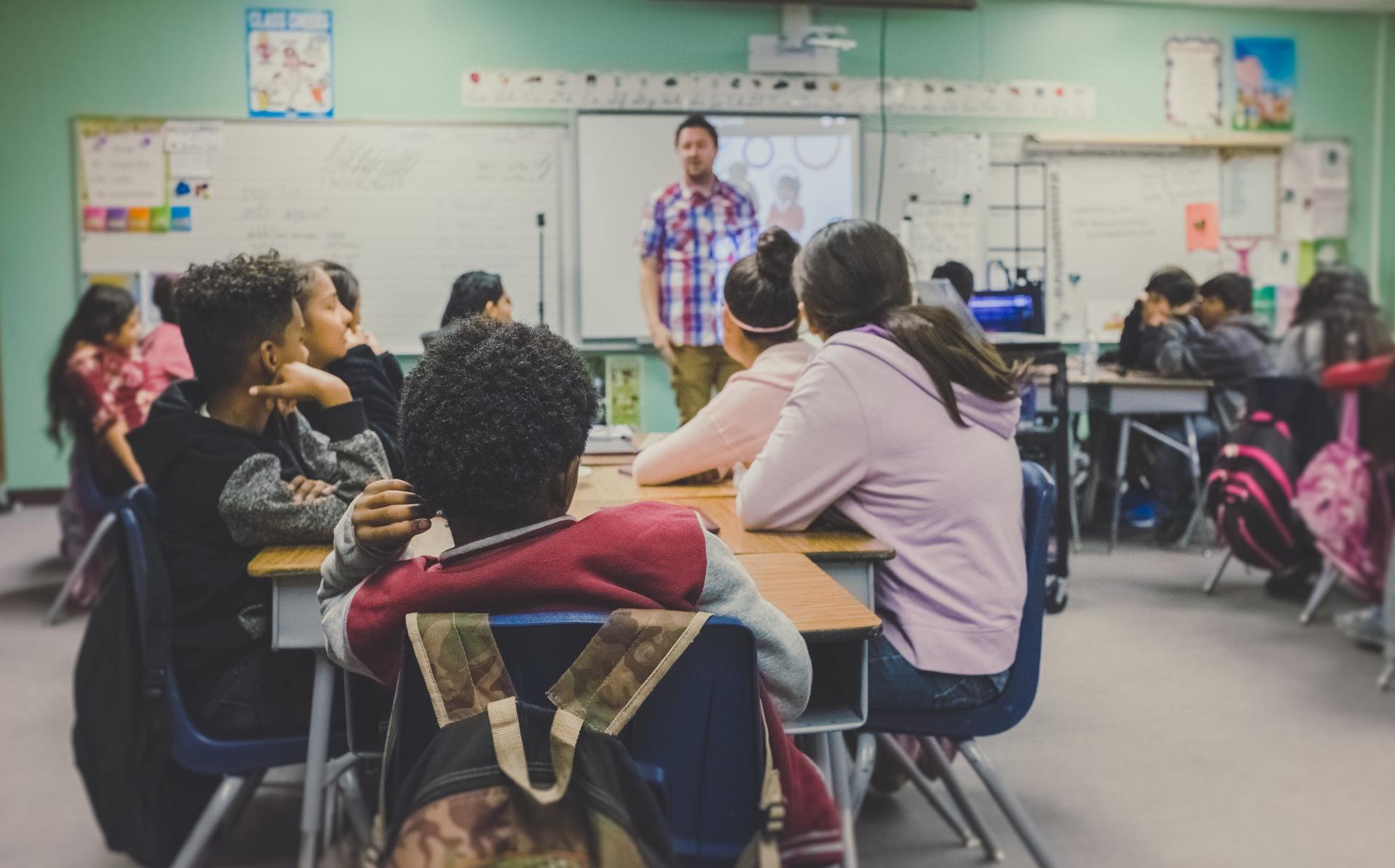 A group of children are sitting at desks in a classroom listening to a teacher.