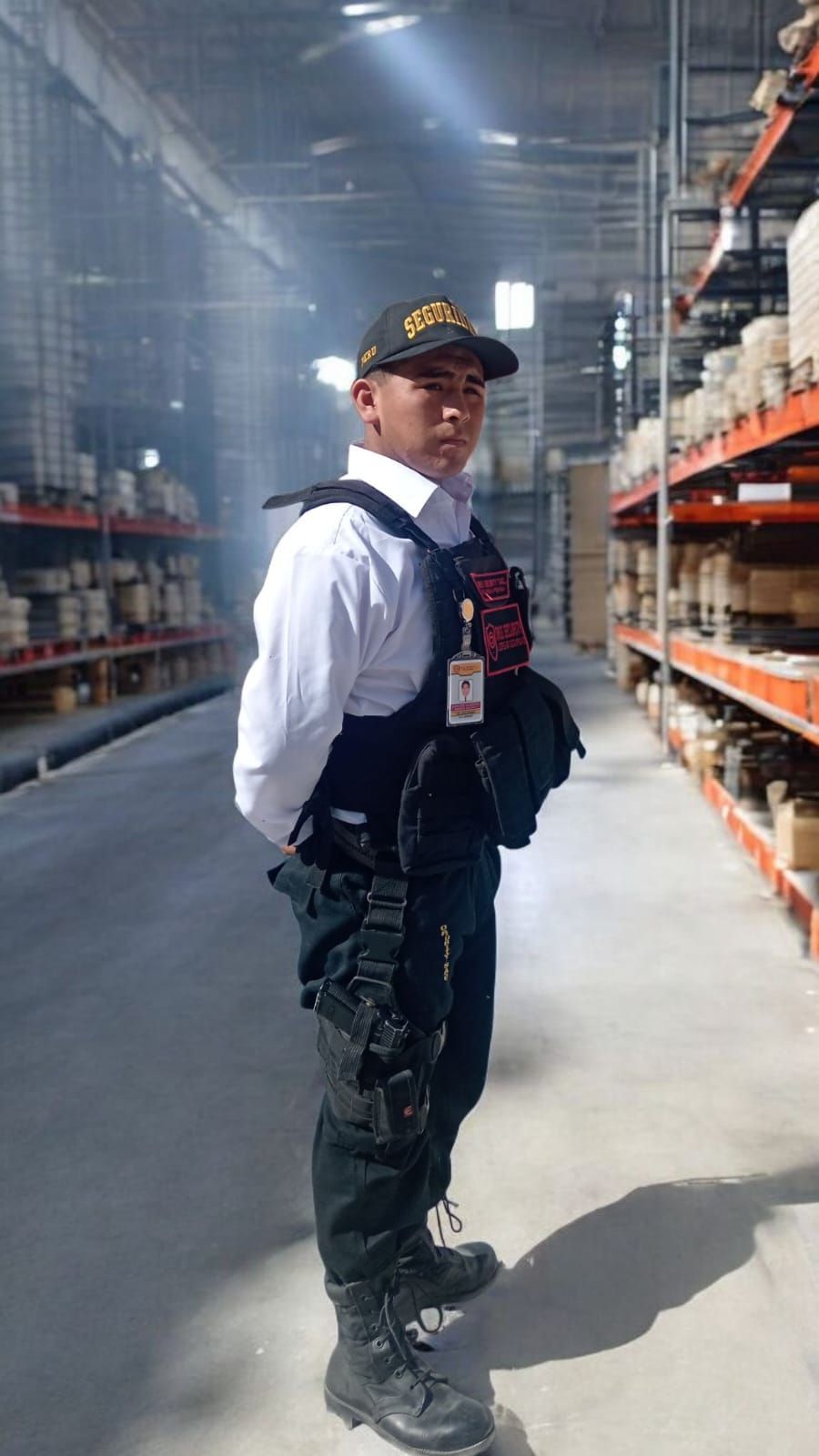 Un hombre con equipo táctico junto a una ambulancia, sosteniendo un rifle. Al aire libre, en un ambiente polvoriento.