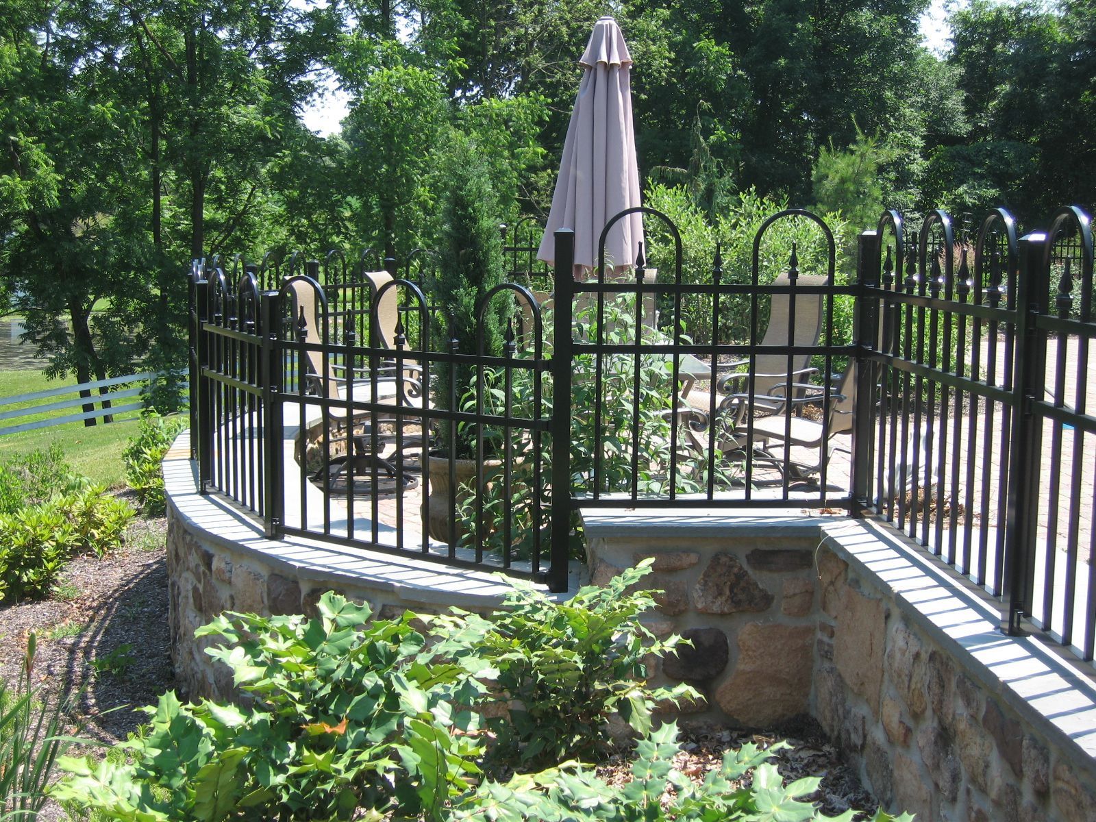 Black metal fence atop a stone wall, surrounding a patio with an umbrella and garden.
