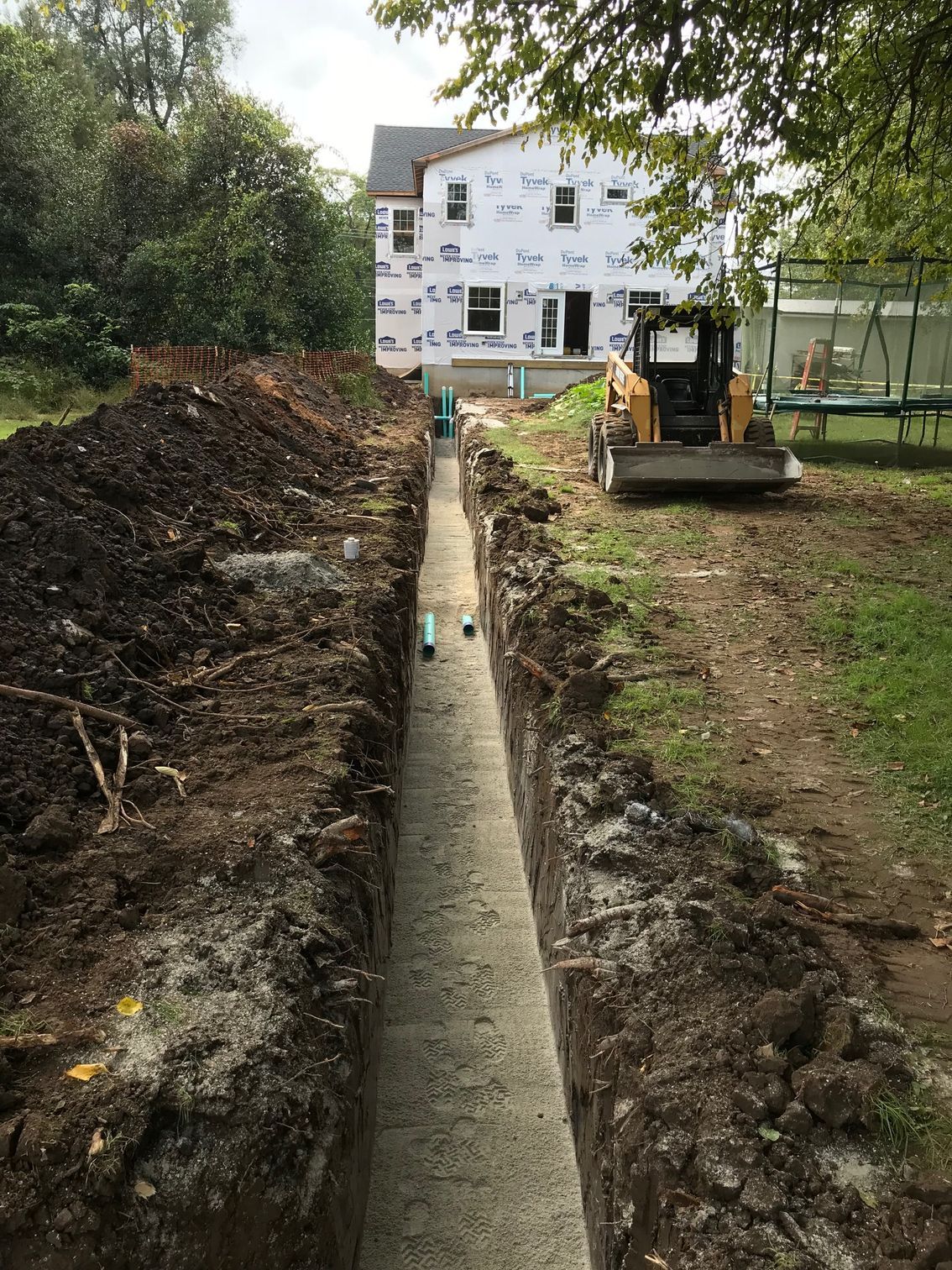 Trench dug in backyard for construction, with exposed piping and heavy machinery. House in background.