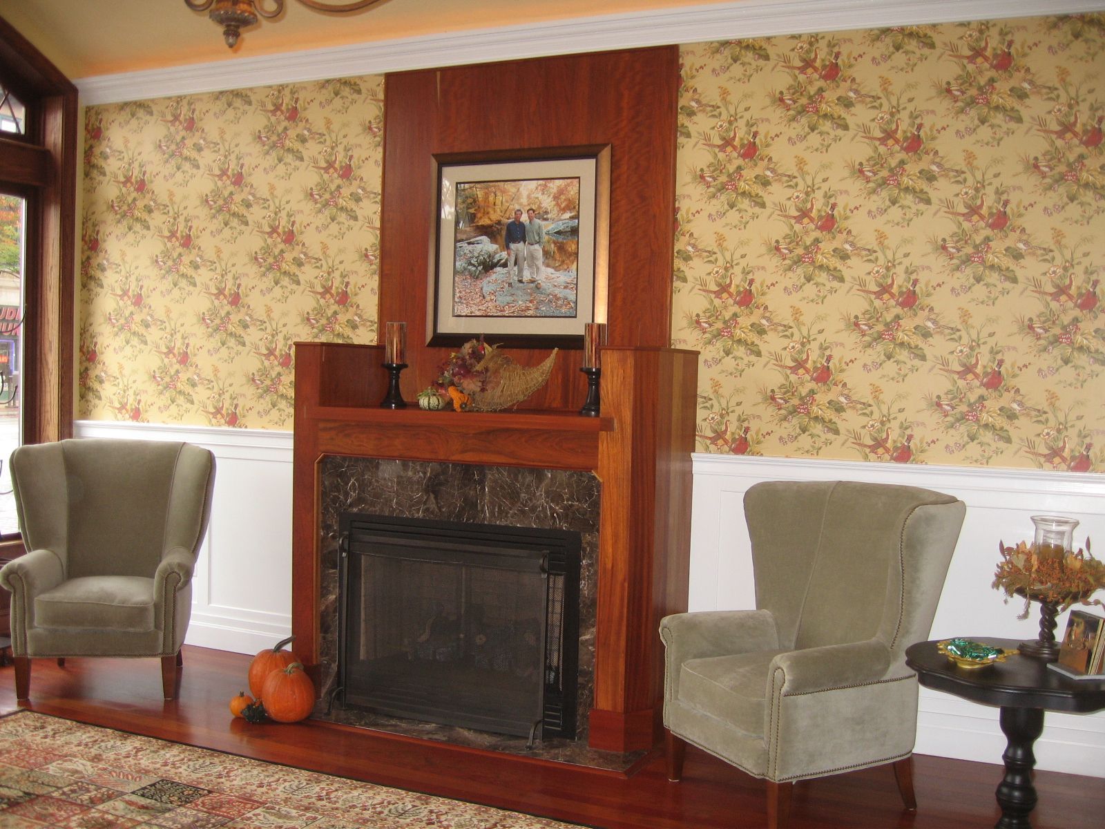 Fireplace flanked by two armchairs, floral wallpaper, wood trim.
