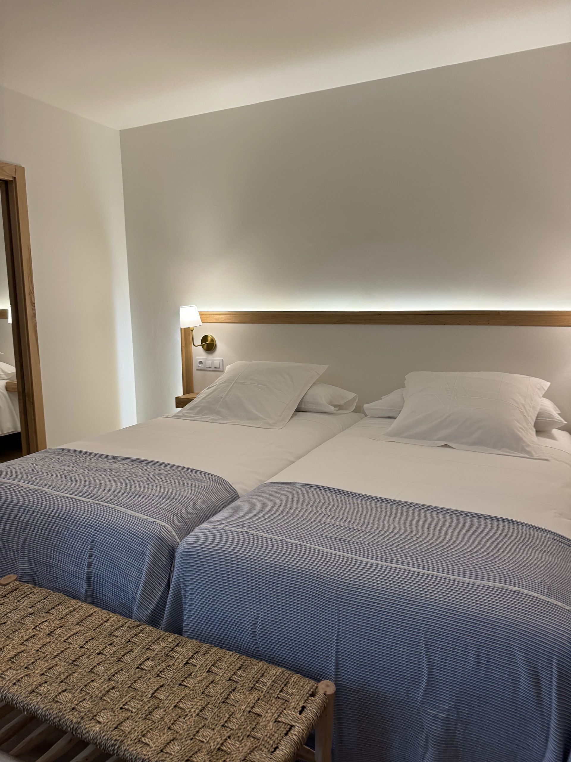 Una habitación de hotel con dos camas individuales con ropa de cama blanca, mantas estampadas en azul y un banco tejido.