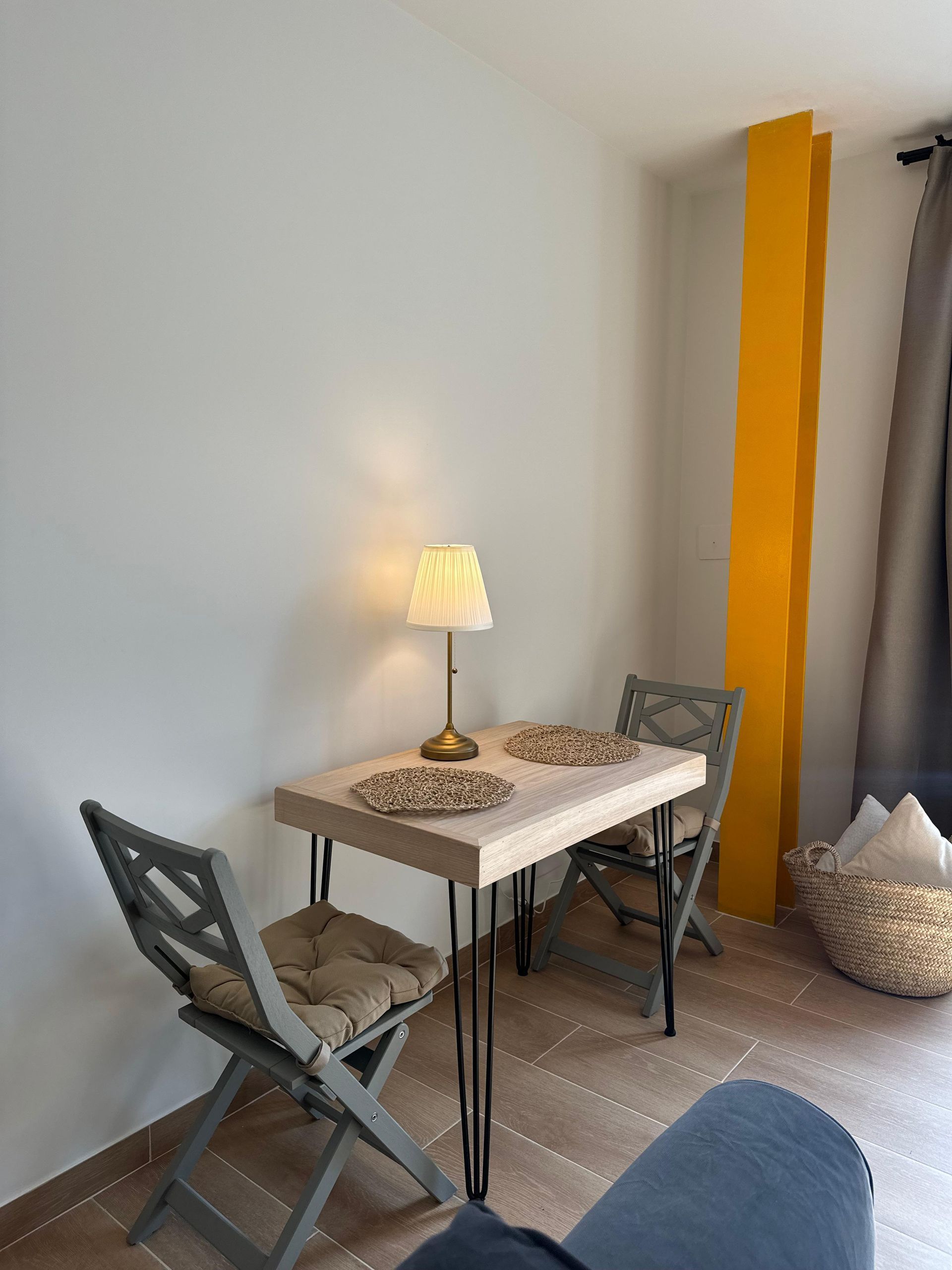 Una pequeña mesa de comedor con dos sillas, lámpara y arte de pared amarillo en una habitación llena de luz.