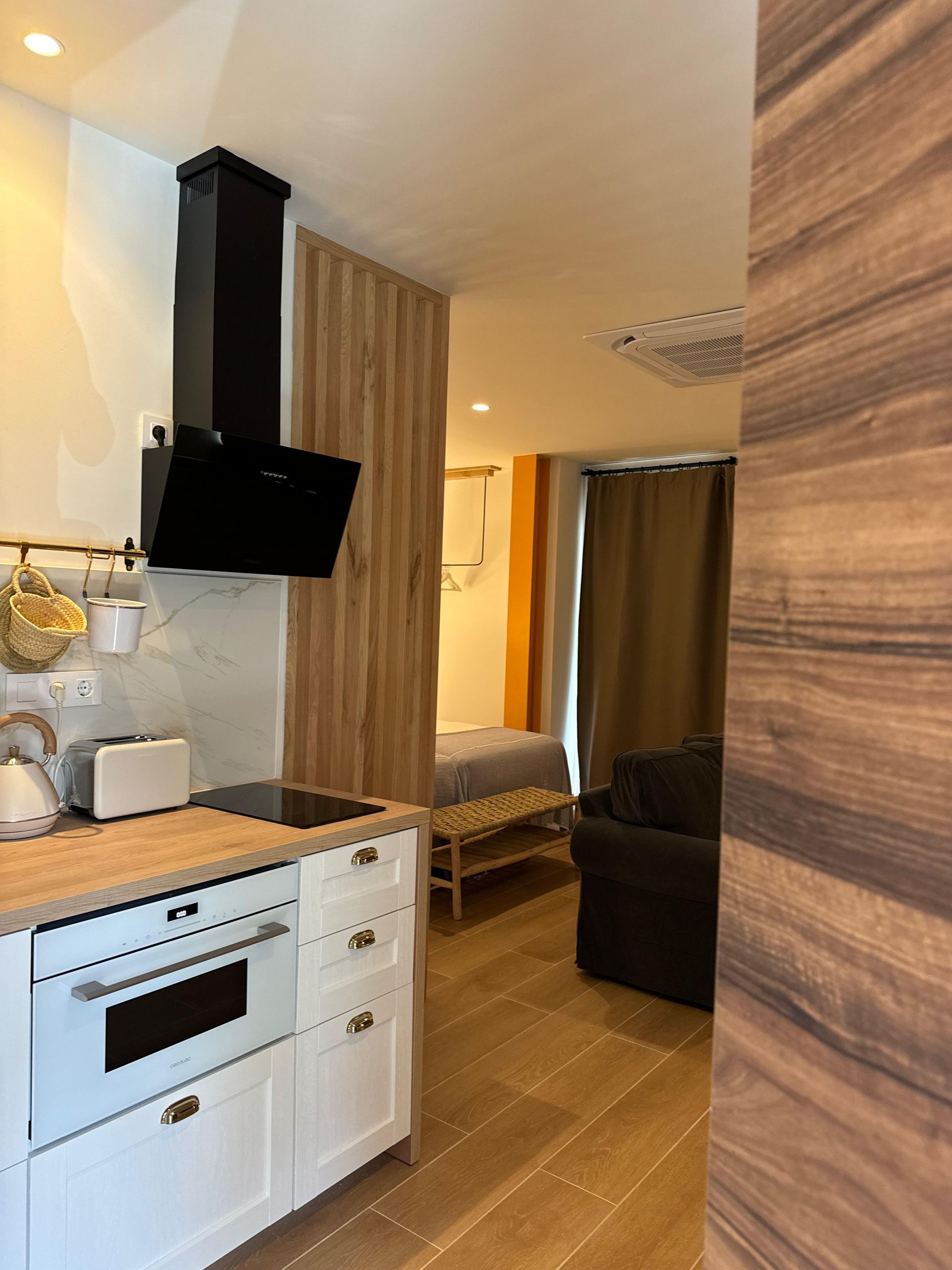 Pequeña cocina con gabinetes blancos, campana extractora negra, detalles de madera, que conduce a un dormitorio con cama