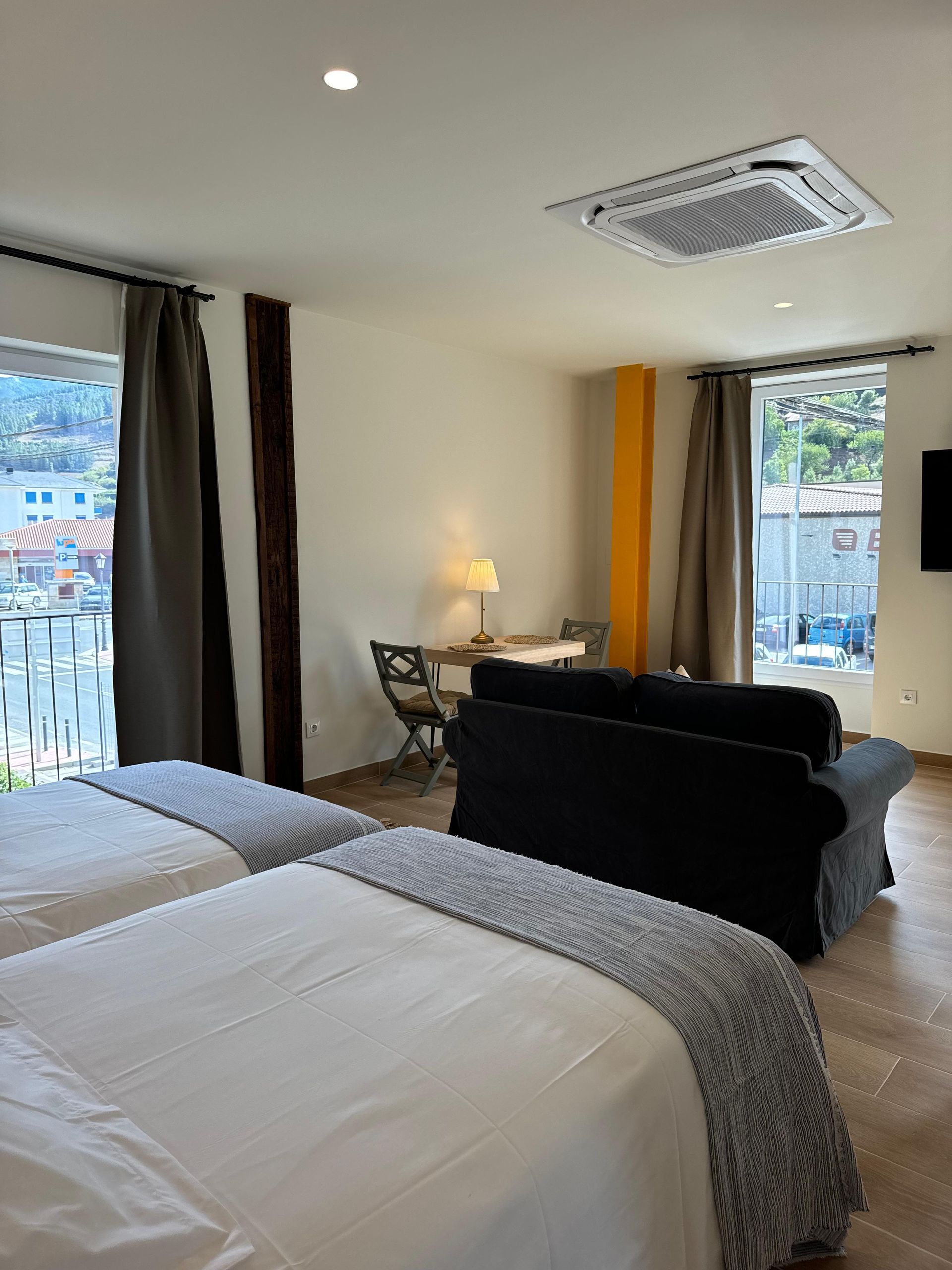 Dormitorio con dos camas, sofá, mesa, ventanas con vistas a la ciudad y una pared de acento de color amarillo brillante.