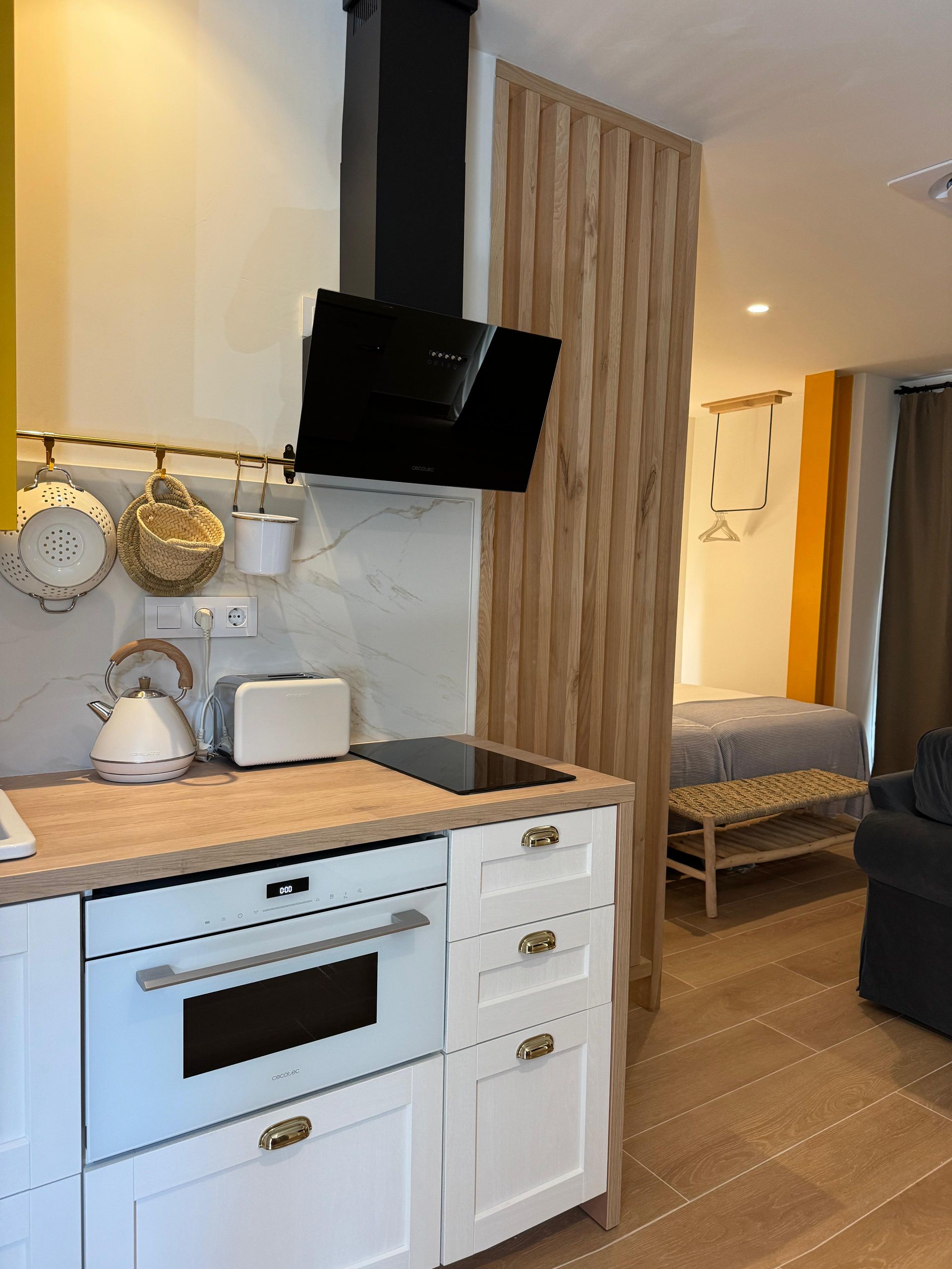 Pequeña cocina con muebles blancos, horno y campana negra, con vista a un dormitorio separado por paneles de madera.