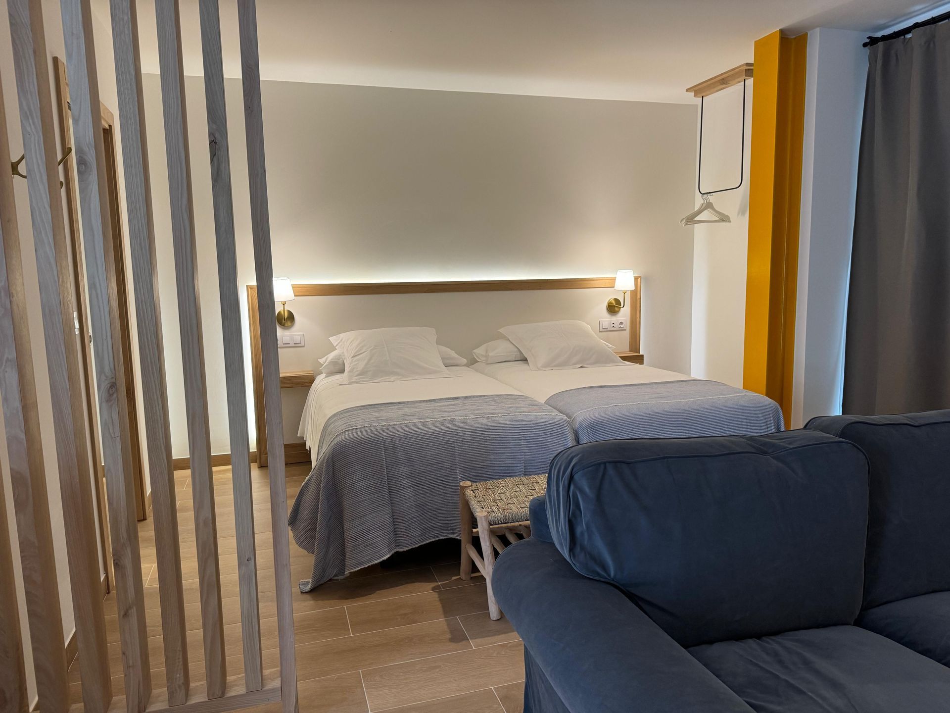 Dormitorio con dos camas, sofá azul, detalles en madera clara y pared amarilla.