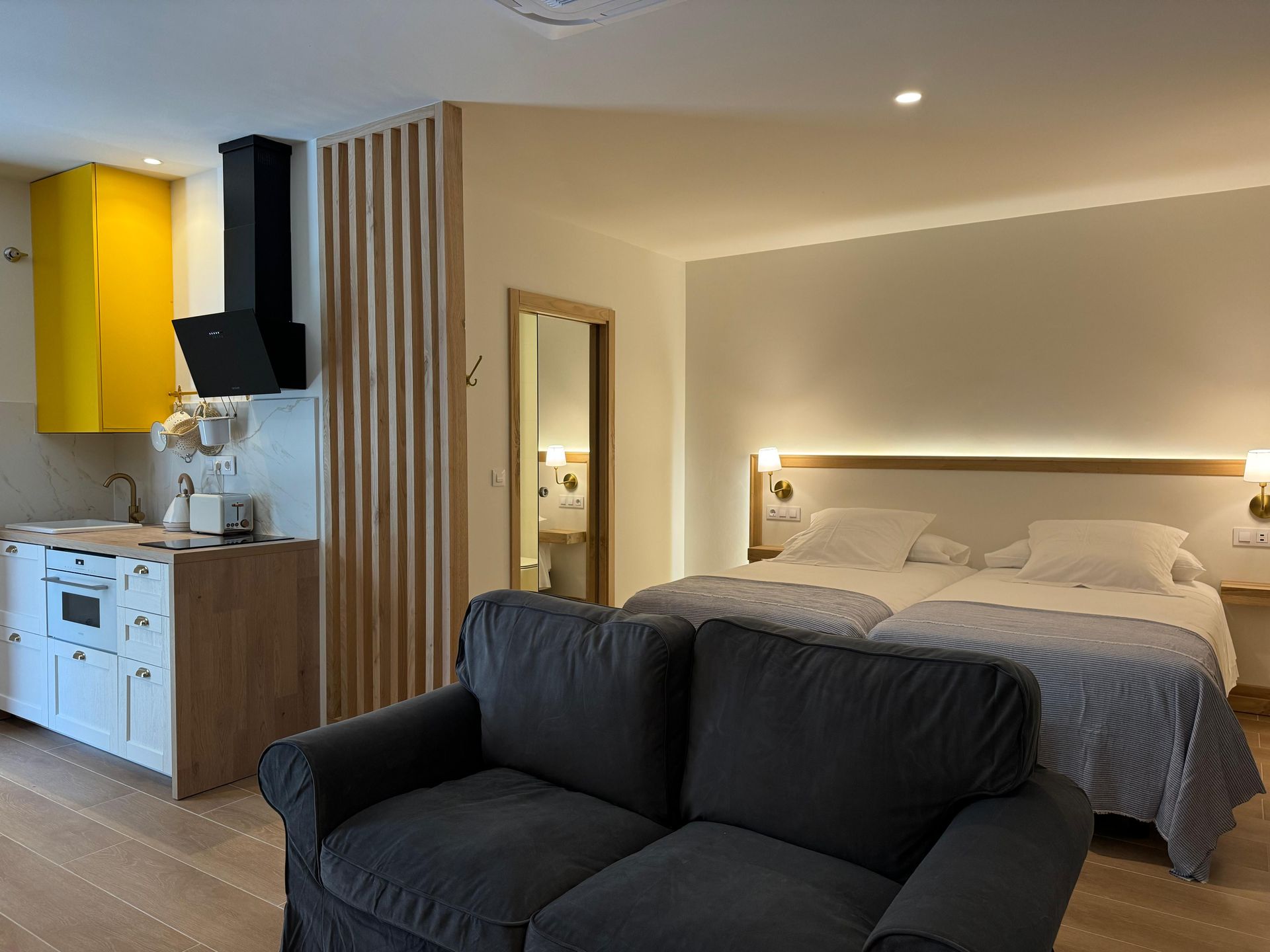 Acogedor apartamento tipo estudio: cama, sofá, cocina americana con armarios amarillos y entrada al baño.