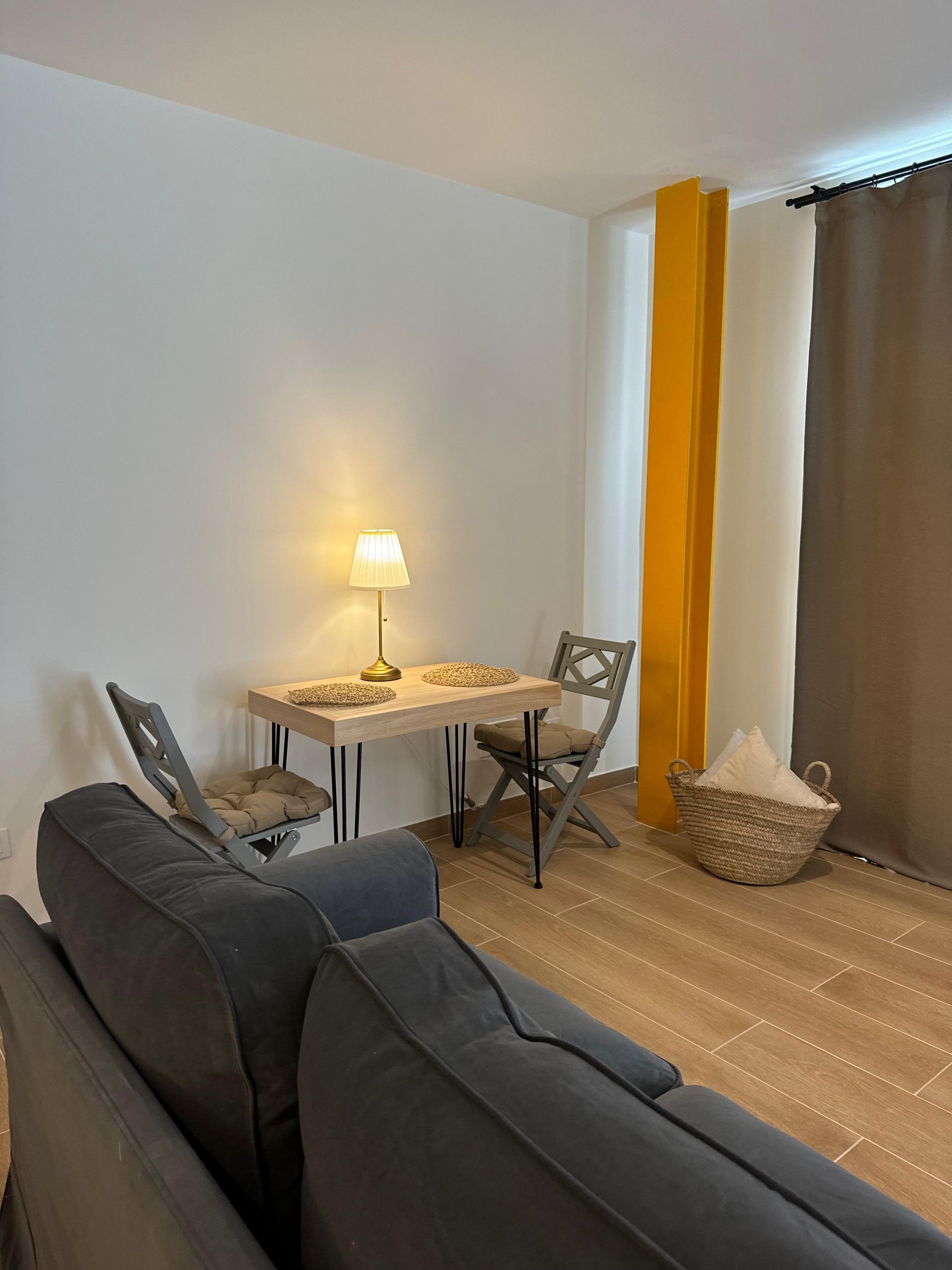 Sala de estar con sofá gris, mesa pequeña con lámpara, dos sillas y viga de soporte amarilla.