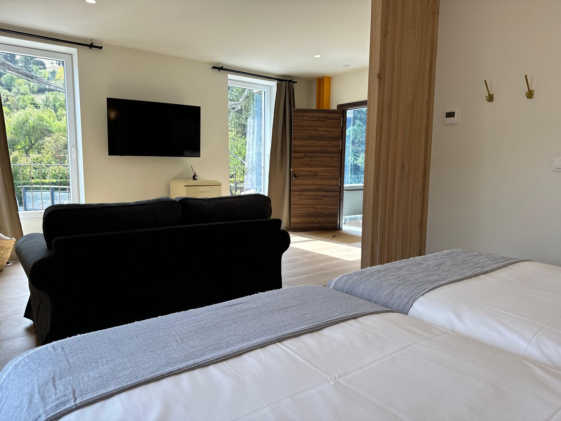 Dormitorio con cama, sofá negro, TV y acceso al balcón. La luz natural ilumina el espacio.
