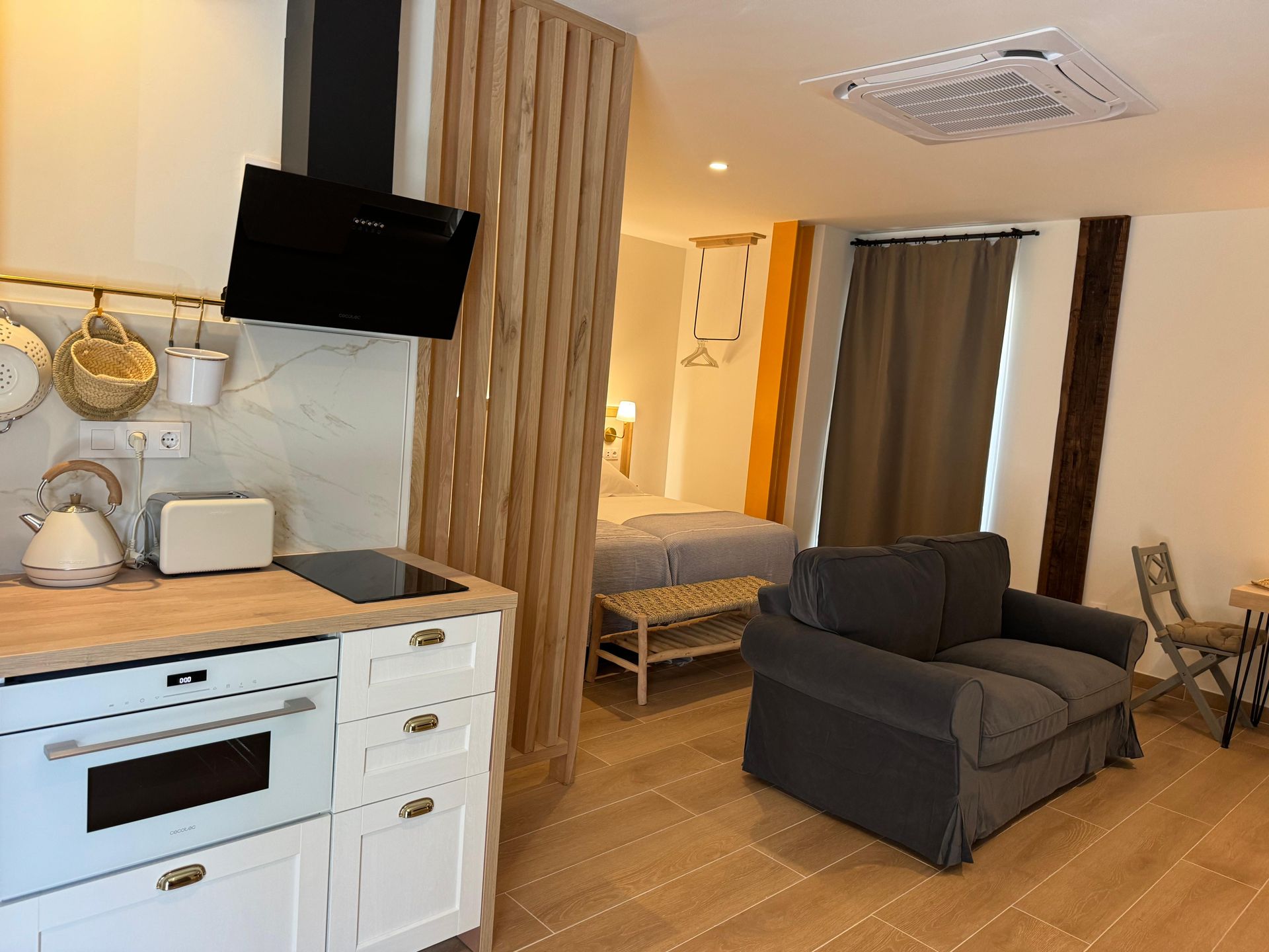 Interior de pequeño apartamento con cocina americana, zona de estar y una cama detrás de una mampara de madera.
