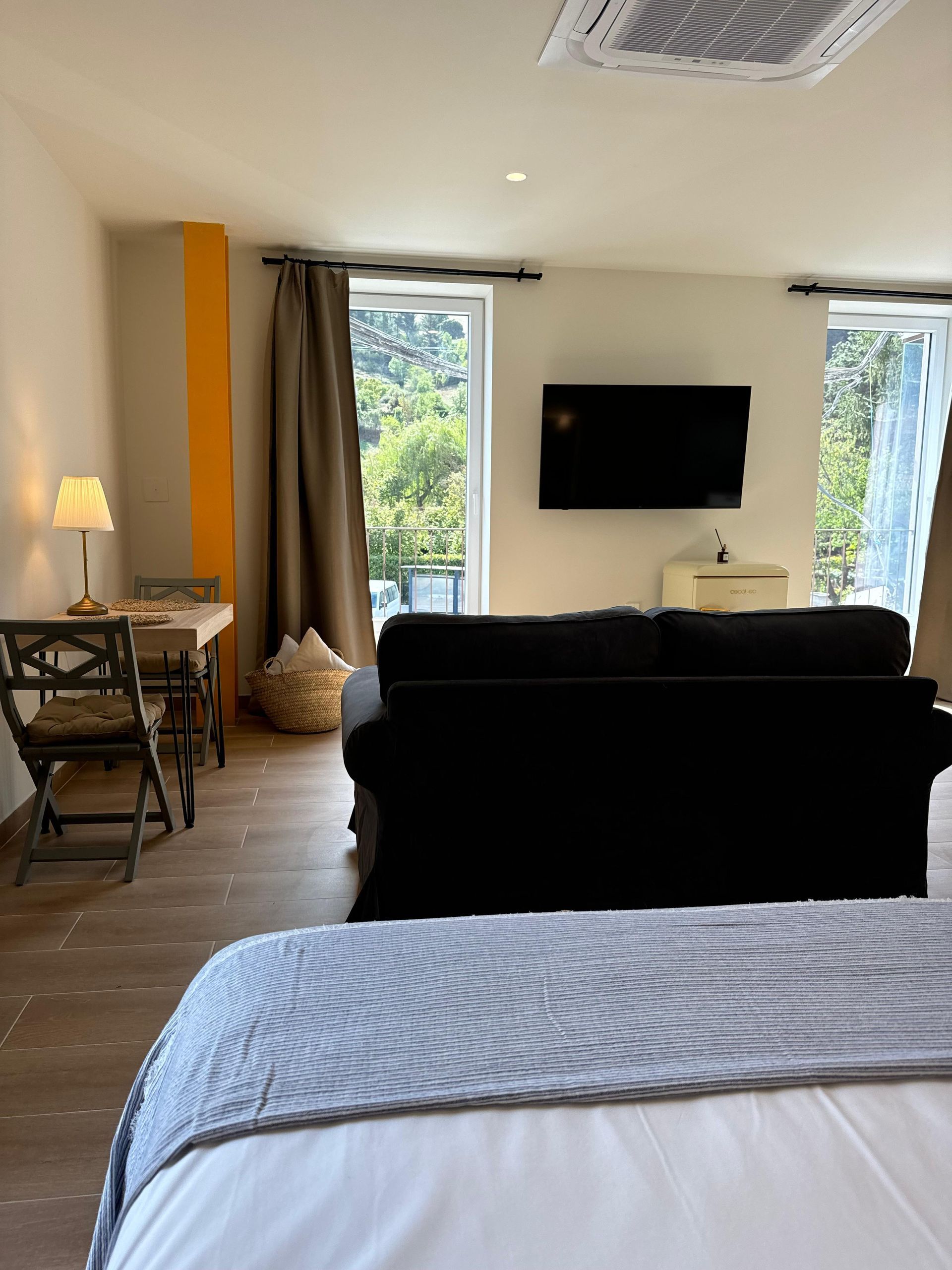Interior de un estudio con cama, sofá, mesa de comedor y televisión. Las ventanas ofrecen vistas a los árboles.