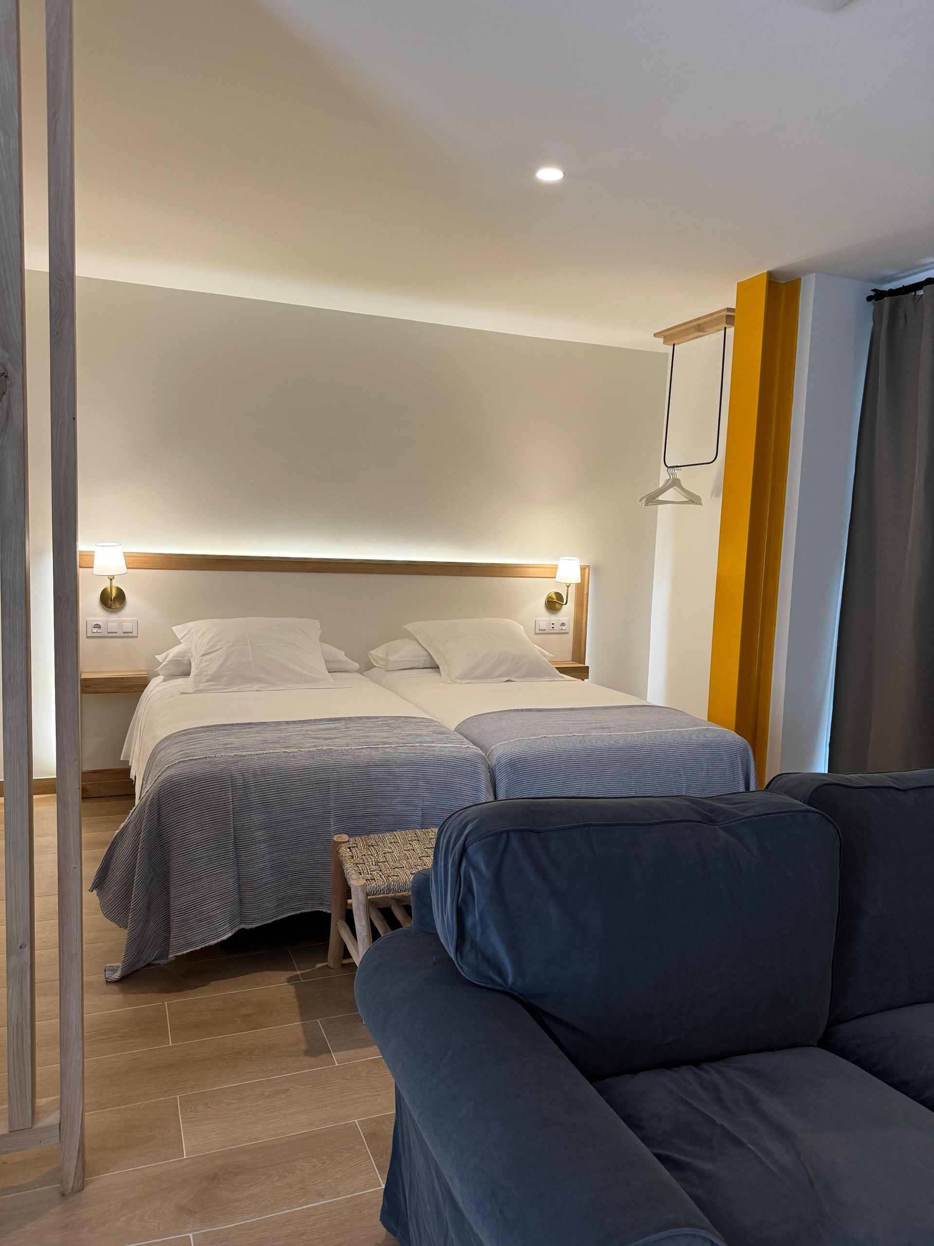 Dormitorio con dos camas individuales, ropa de cama gris y blanca, sofá en primer plano. Detalles en beige y amarillo.