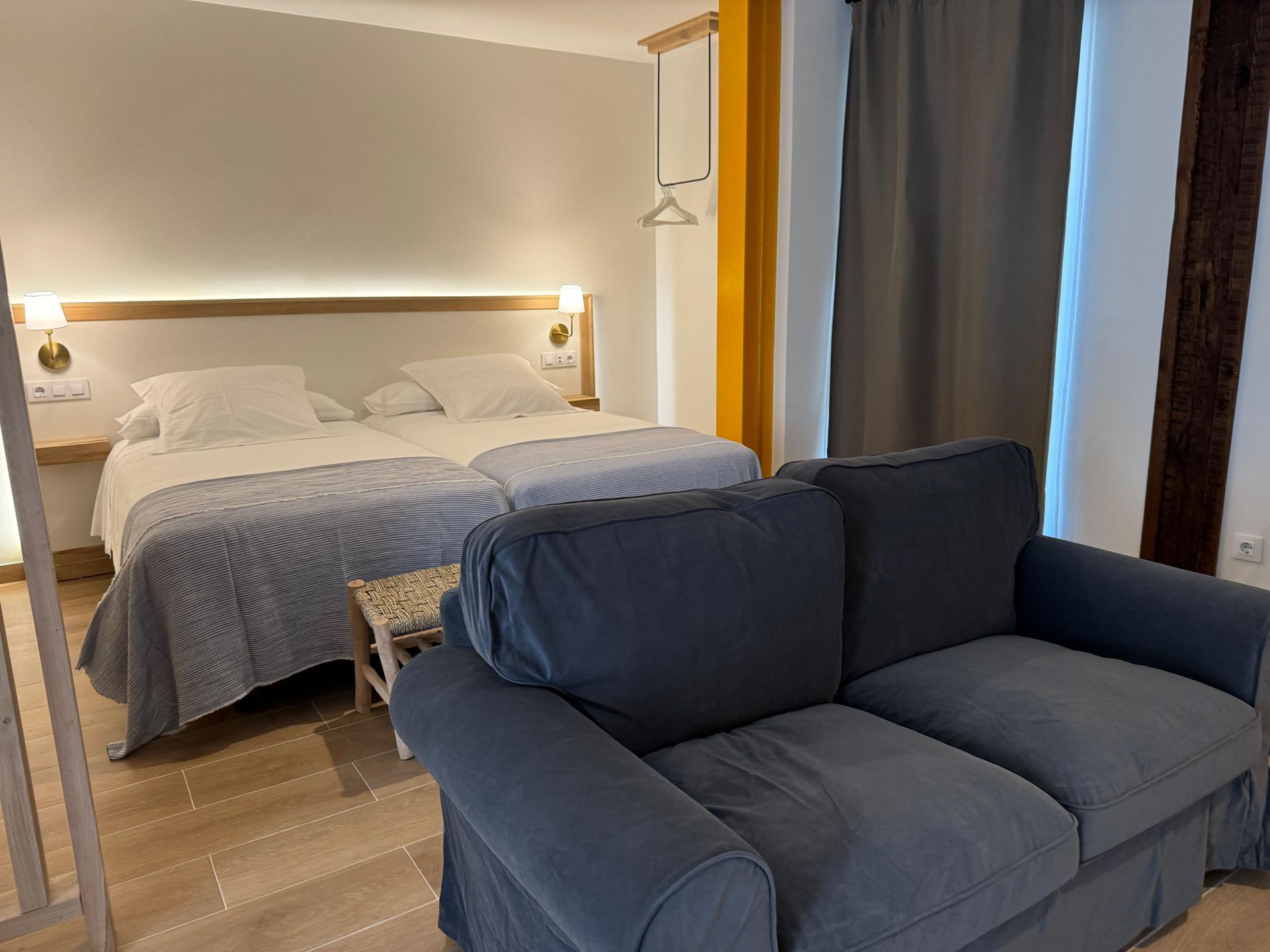 Una habitación de hotel con dos camas y un sofá azul. Paredes blancas, suelos de madera y colores neutros.