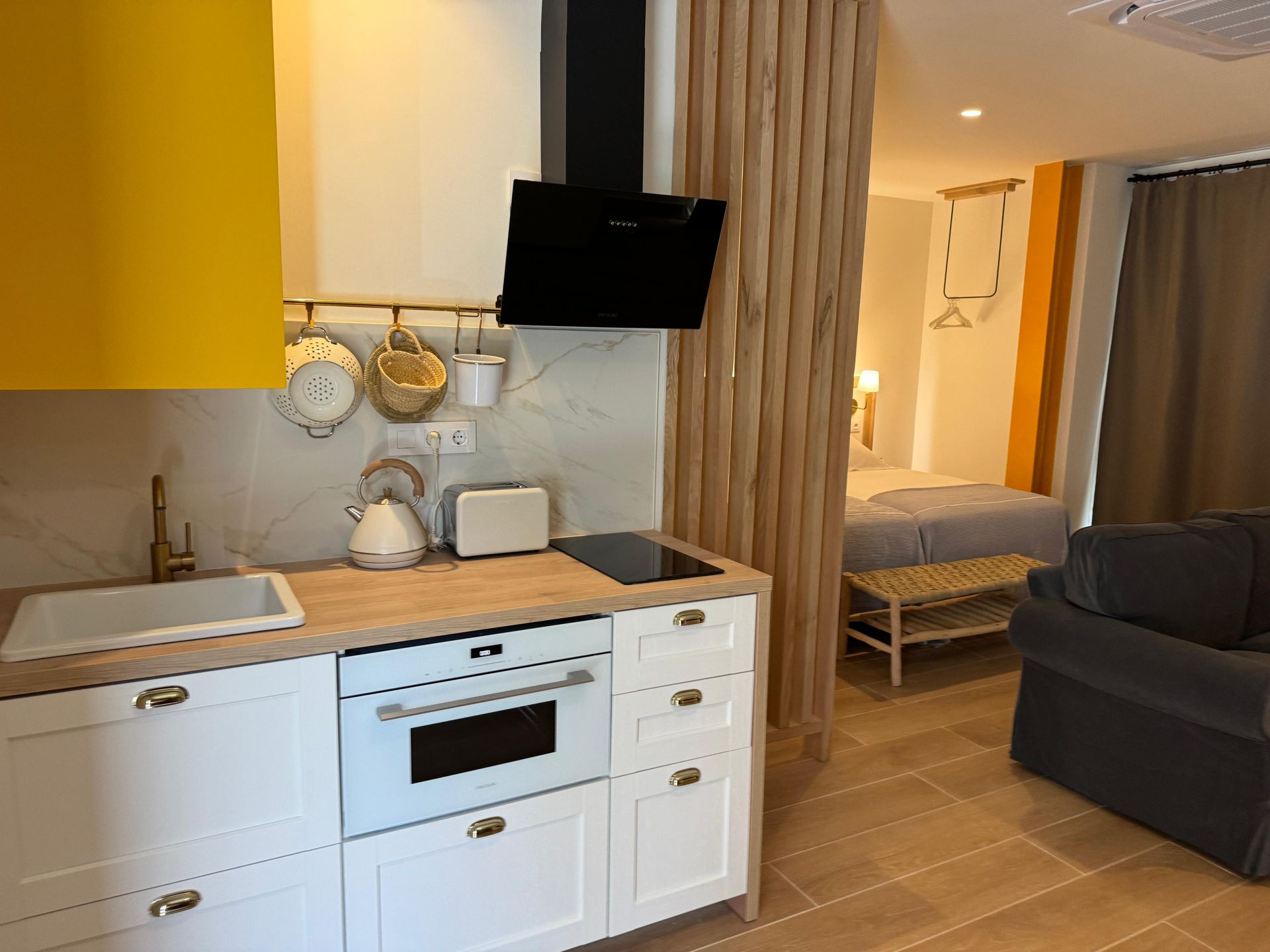Área de cocina con gabinetes blancos, detalles en amarillo y vista a un dormitorio con cama y banco.