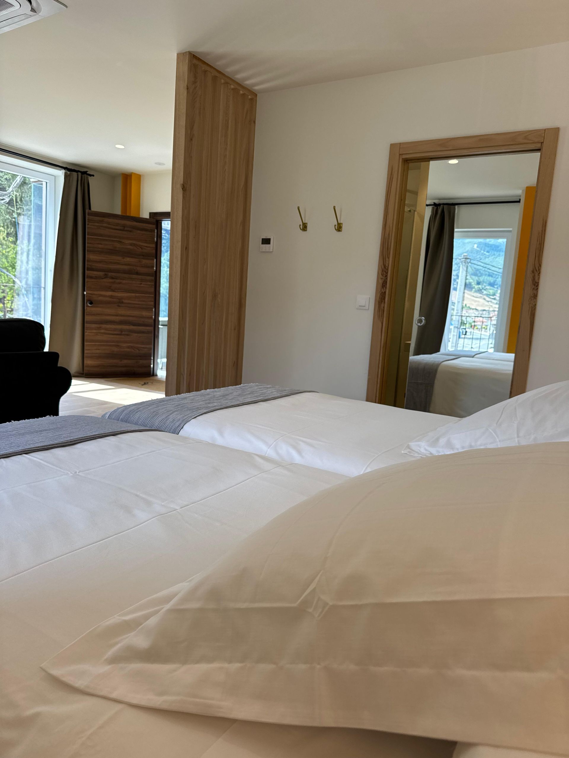 Dormitorio con ropa de cama blanca, espejo grande, pared con acento de madera y vista al exterior.