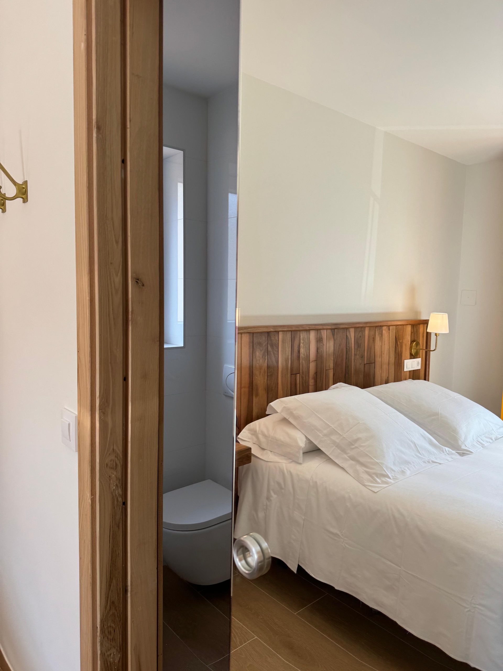 Puerta del dormitorio con vista a un baño y una cama con sábanas blancas. Molduras de madera y paredes claras.