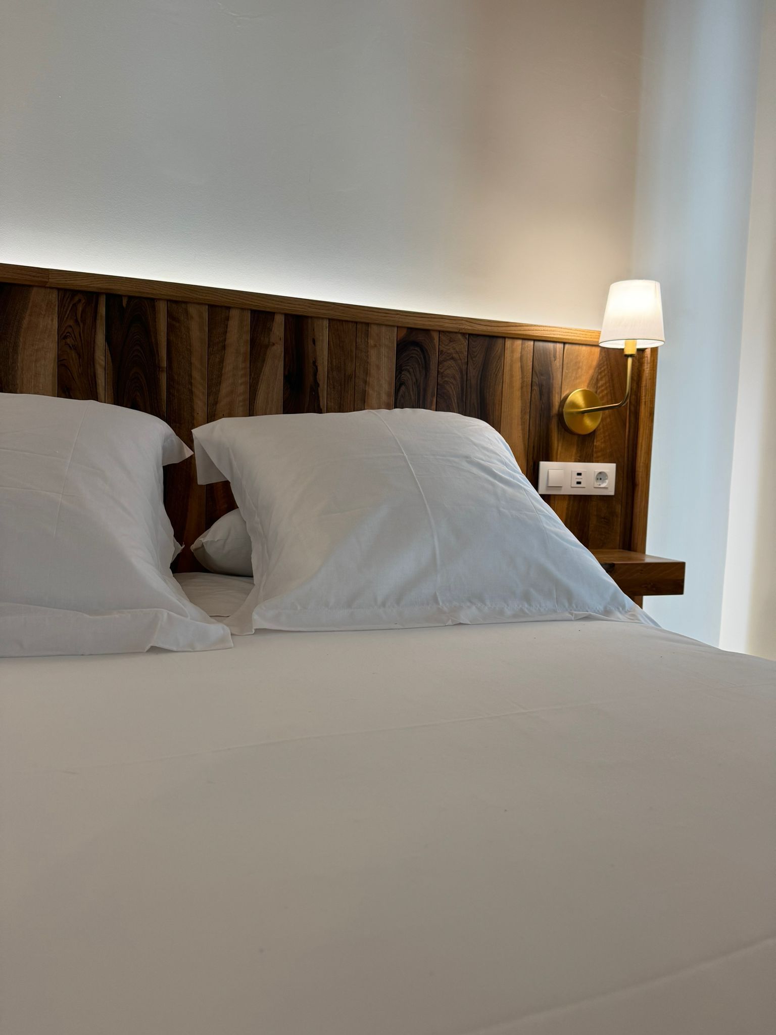 Cama con sábanas blancas, cabecero de madera y lámpara de noche.