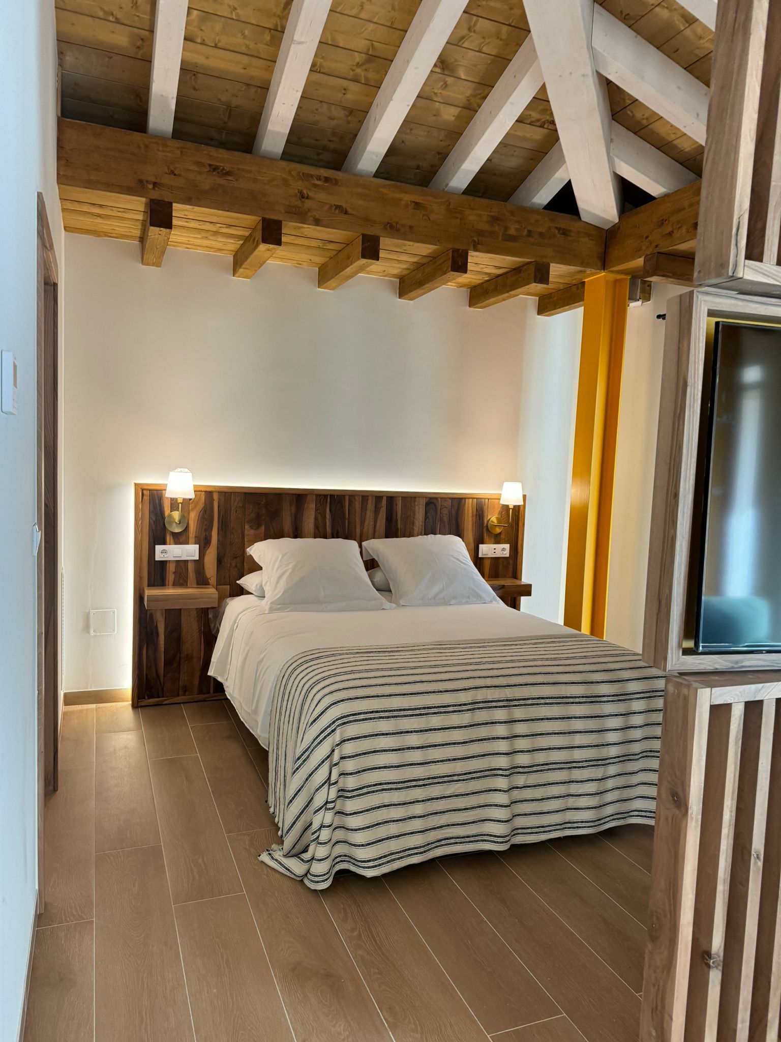 Dormitorio con cabecero, vigas y suelo de madera. Ropa de cama blanca, manta estampada e iluminación cálida.