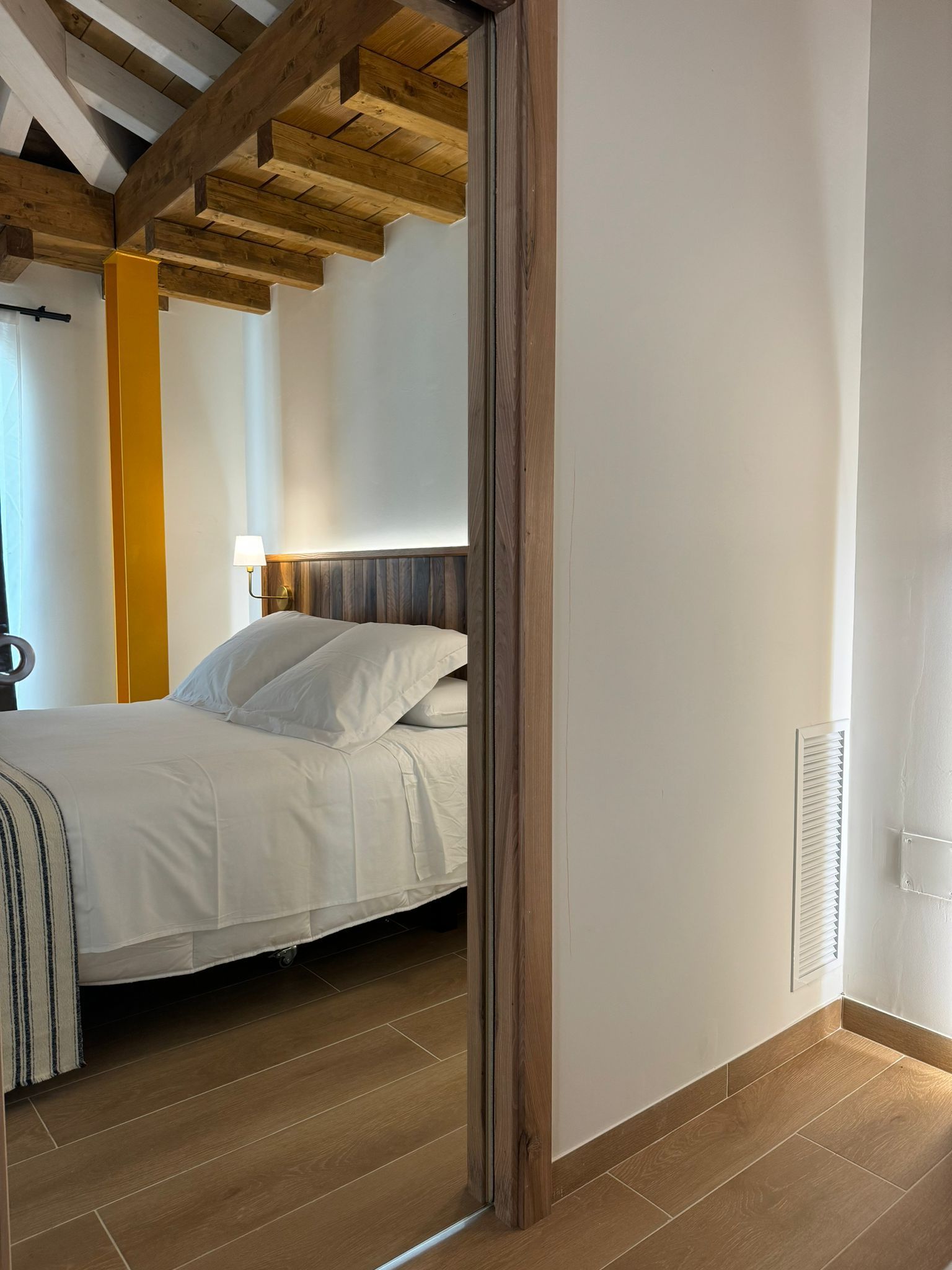 Interior del dormitorio: ropa de cama blanca, vigas de madera, detalles en amarillo, visibles a través de la puerta.