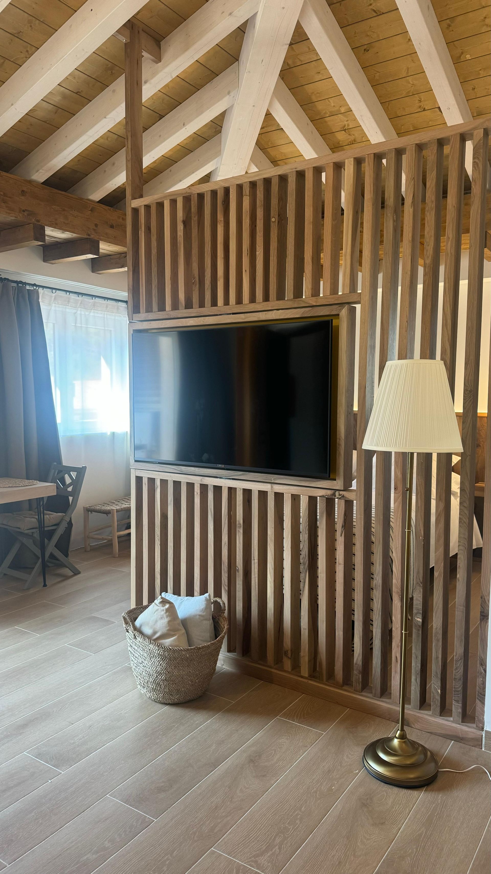 Biombo de listones de madera con TV montada, cesta y lámpara de pie en una habitación con suelo de madera.