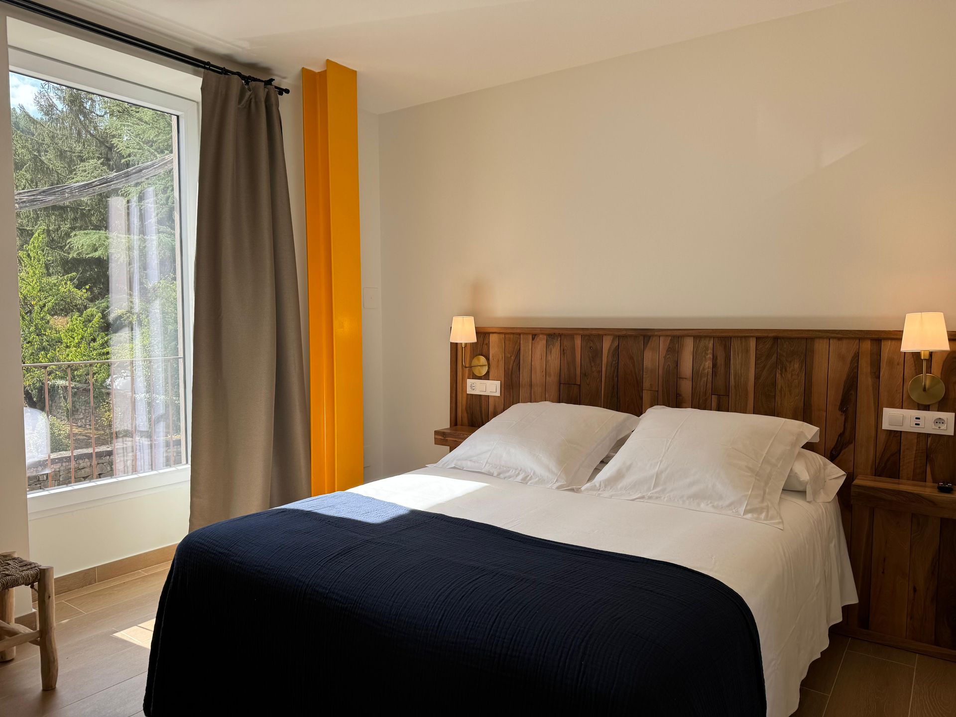 Dormitorio con cama doble, cabecero de madera y ventana. Entra la luz del sol a raudales.