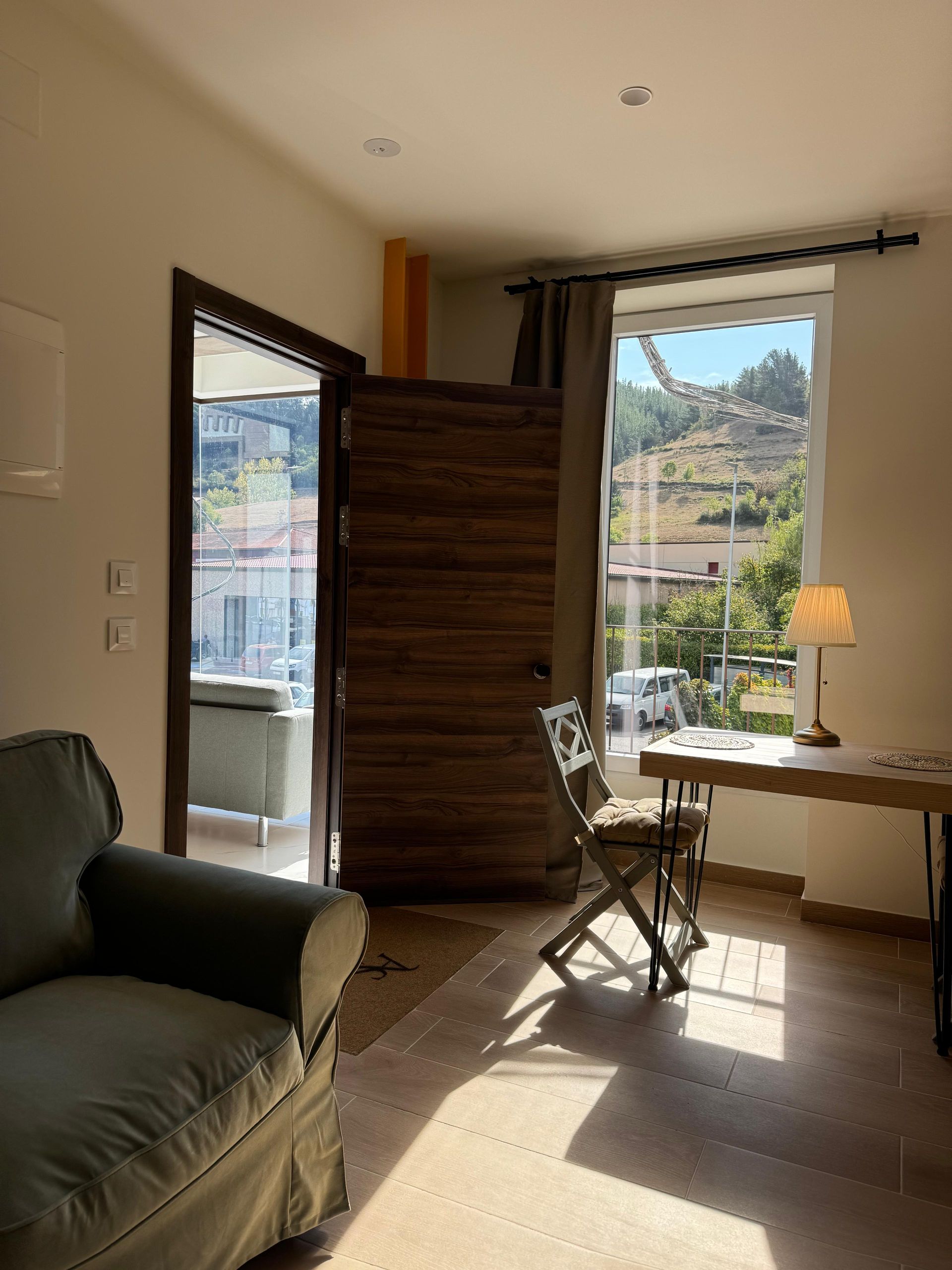  habitación con puerta abierta al balcón, silla junto a la ventana con escritorio , sofá y vista soleada de la colina.