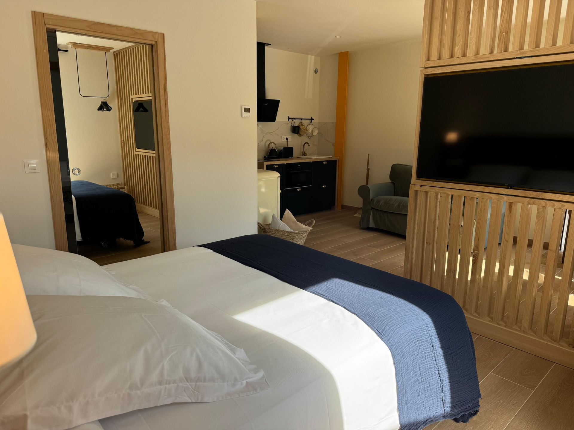 Dormitorio con cama, espejo, cocina y TV; interior iluminado por el sol con detalles en azul, blanco y madera.